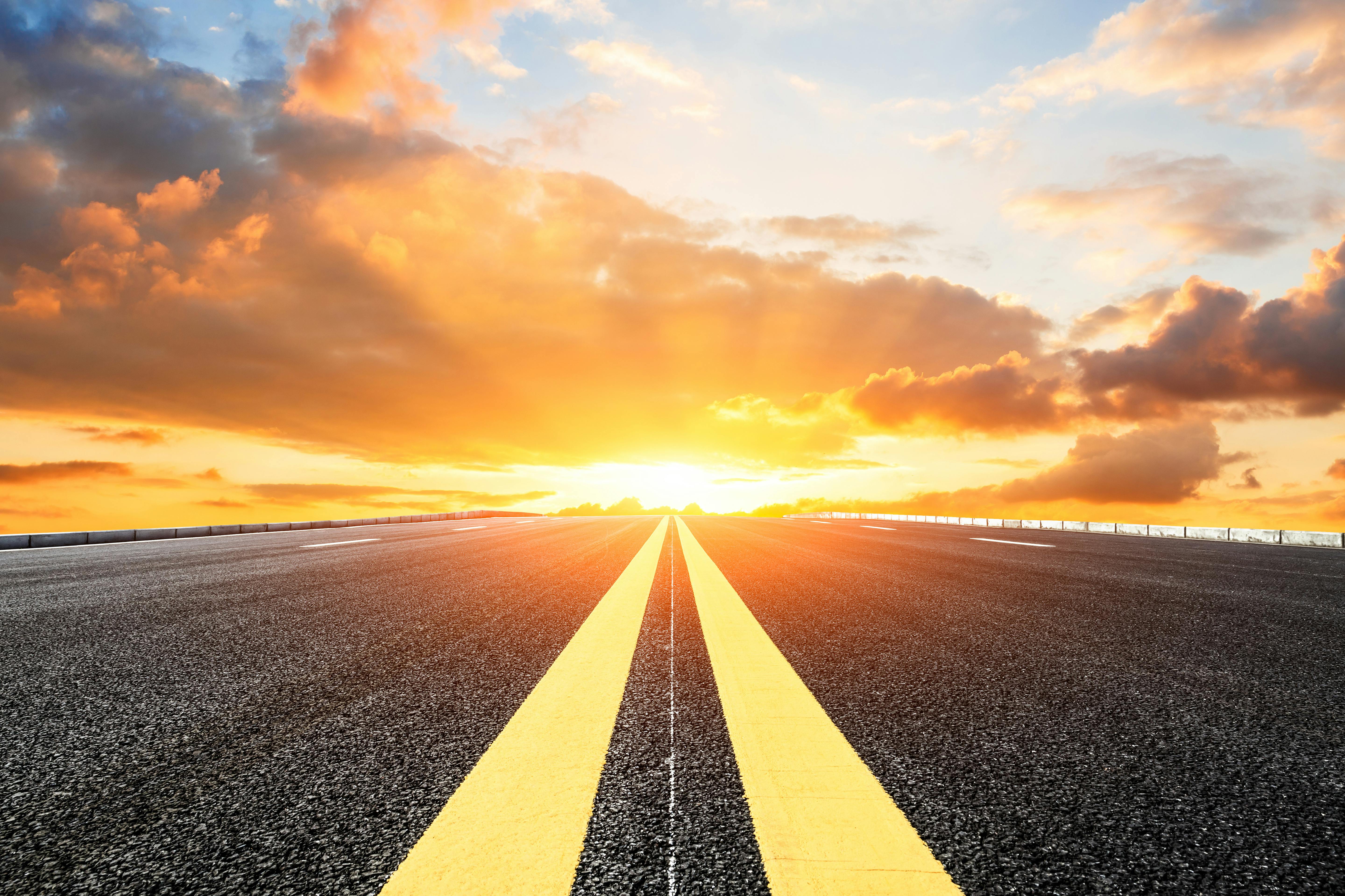 A long, straight road stretches towards the horizon under a dramatic sunset sky.