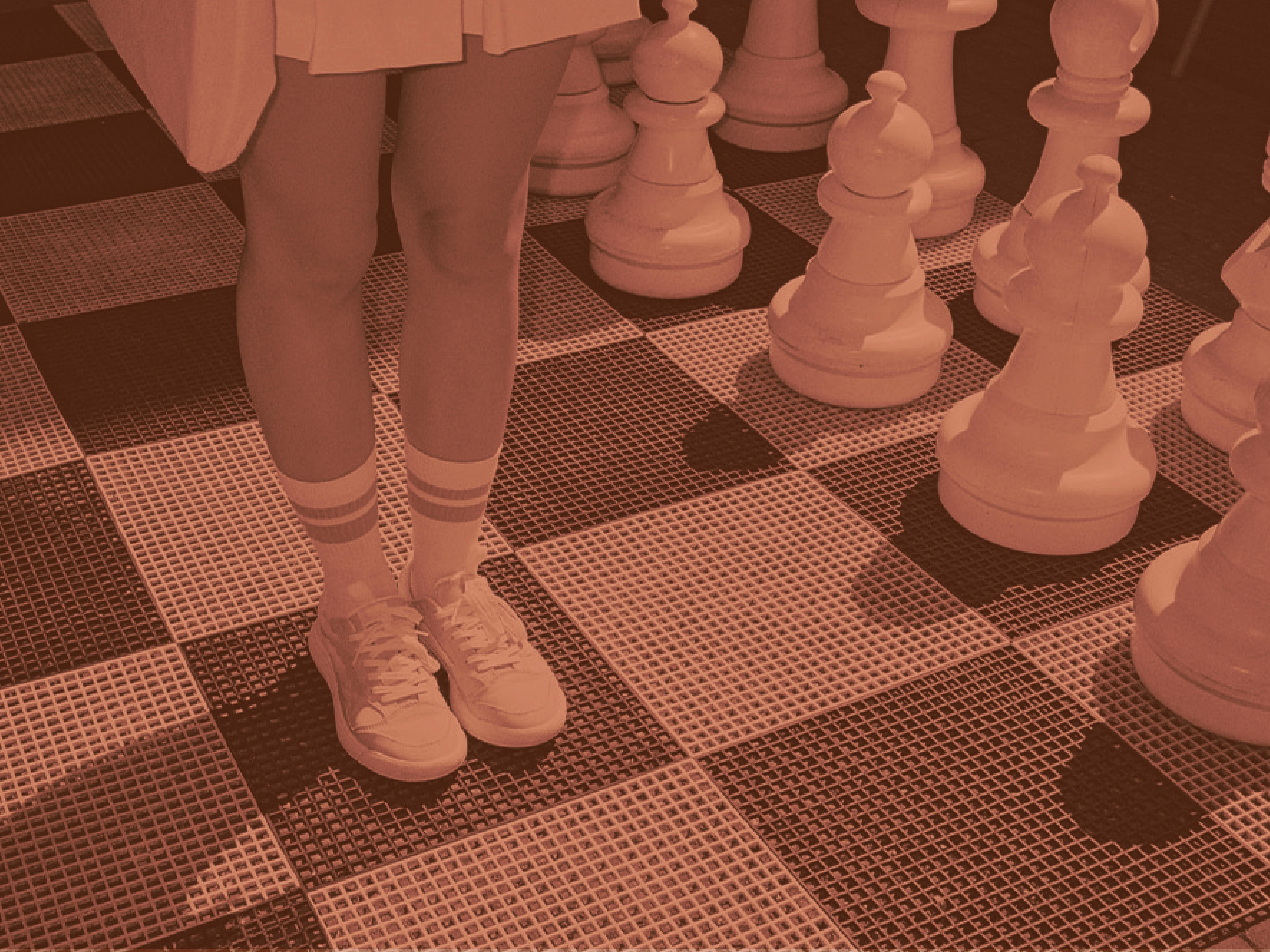 Person standing on a giant outdoor chessboard next to oversized chess pieces, wearing sneakers and striped socks.
