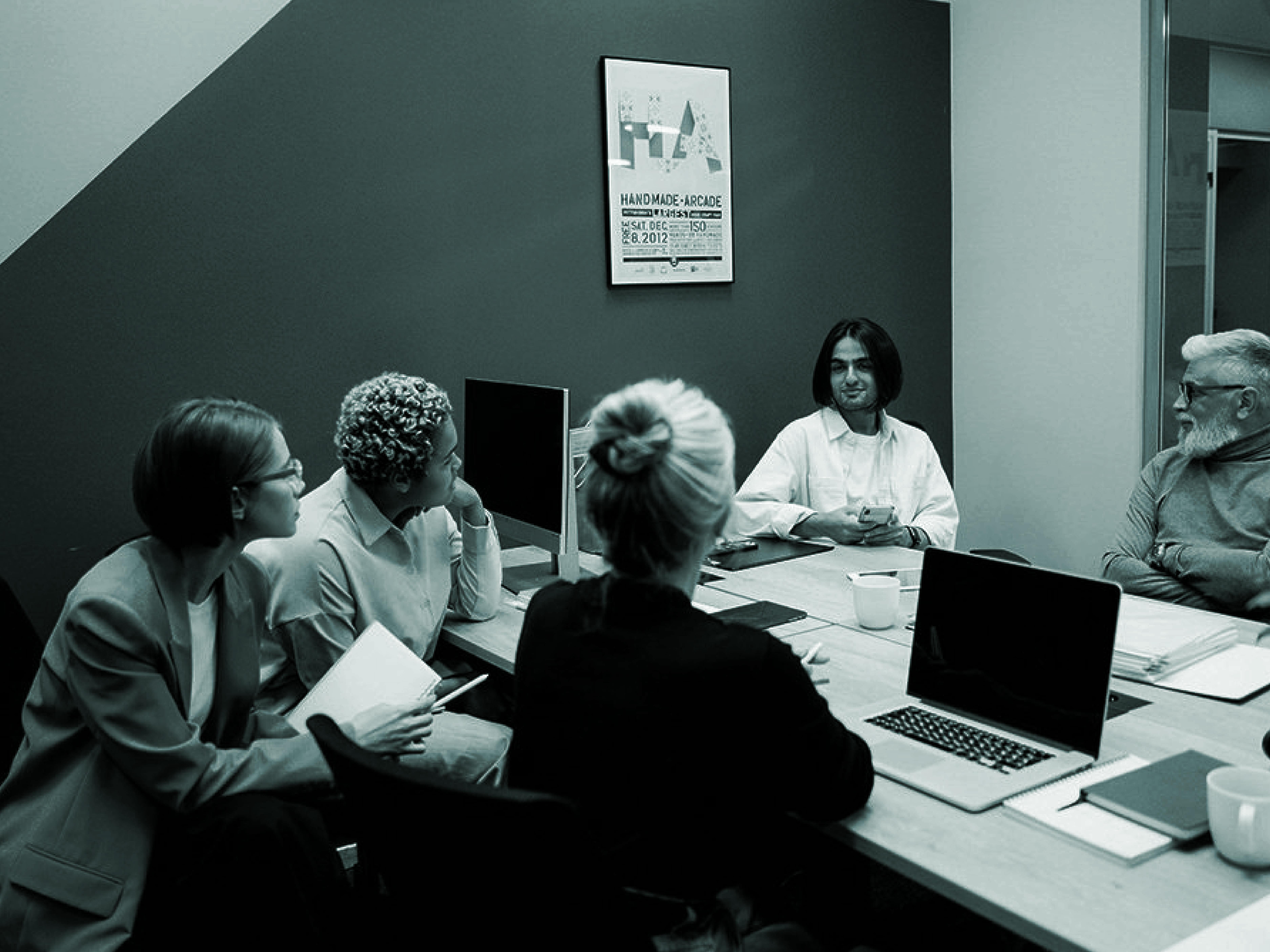A group of five people sitting around a conference table engaged in a meeting, with laptops, notebooks, and coffee cups in front of them.