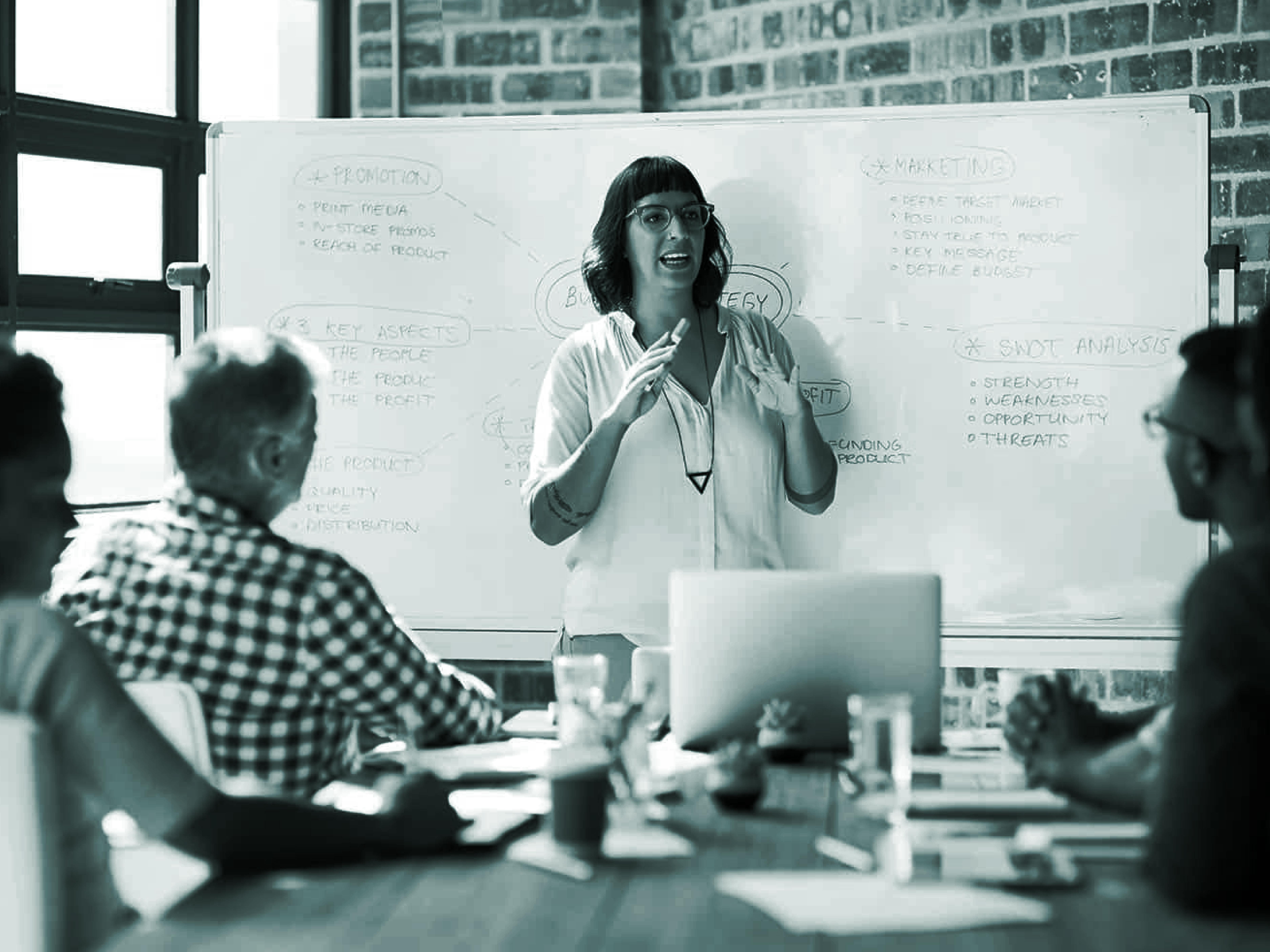 A woman stands and presents in front of a whiteboard filled with strategy notes, while four colleagues seated around a table listen attentively.