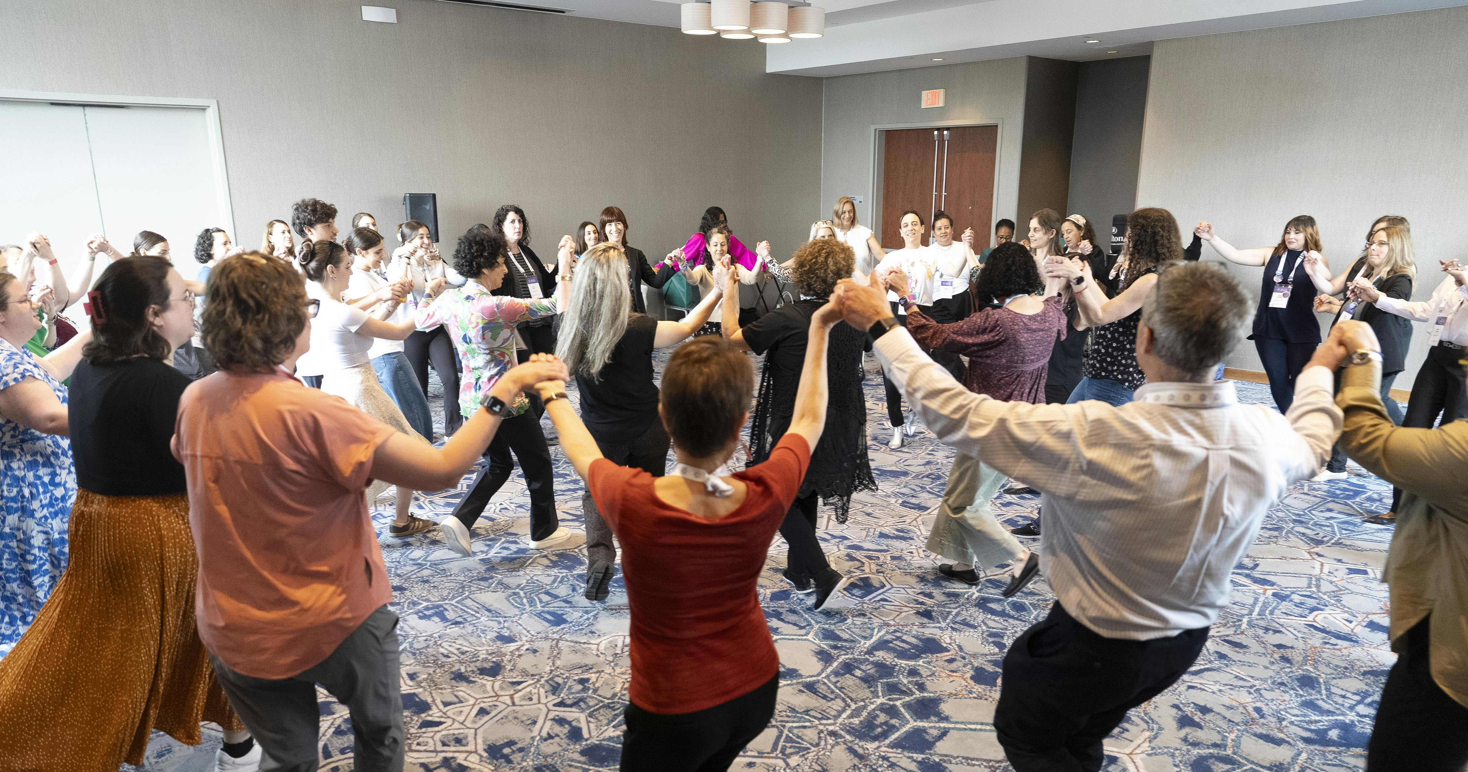 A large group of people stand in a circle holding hands, participating in a traditional group dance during JPro25.