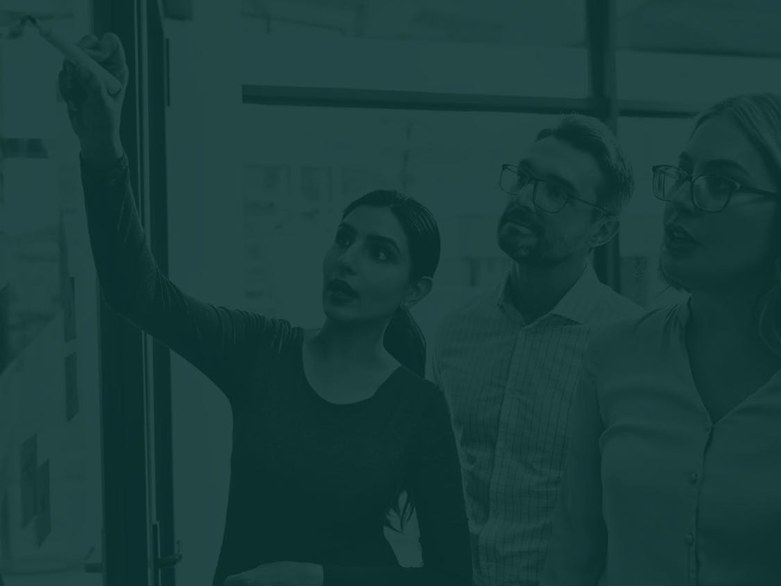 Three people standing together as one points at a board or glass wall during a discussion.