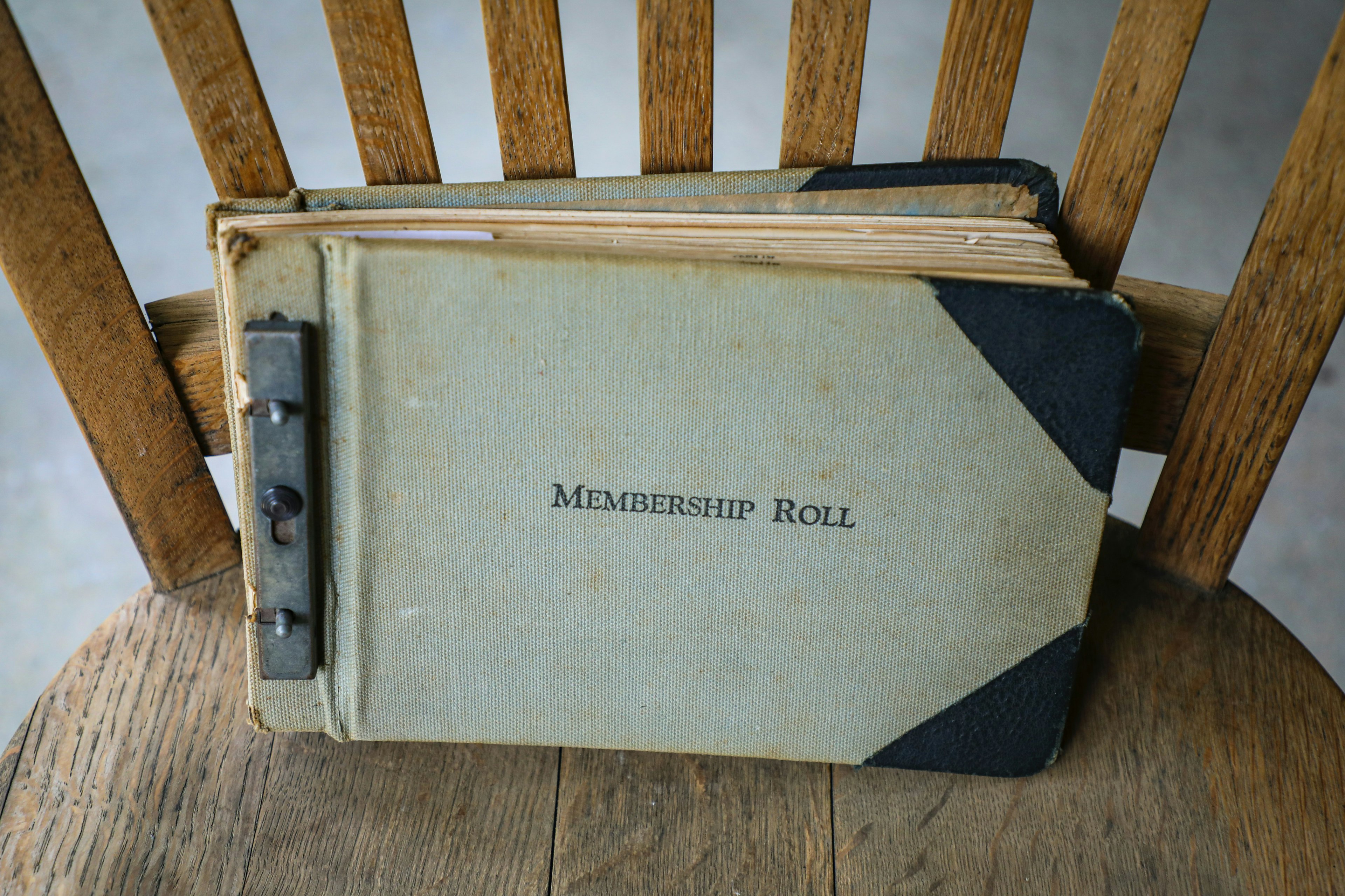 An old and tattered binder with "Membership Roll" embossed on the front, leaning against the back of a wooden chair.