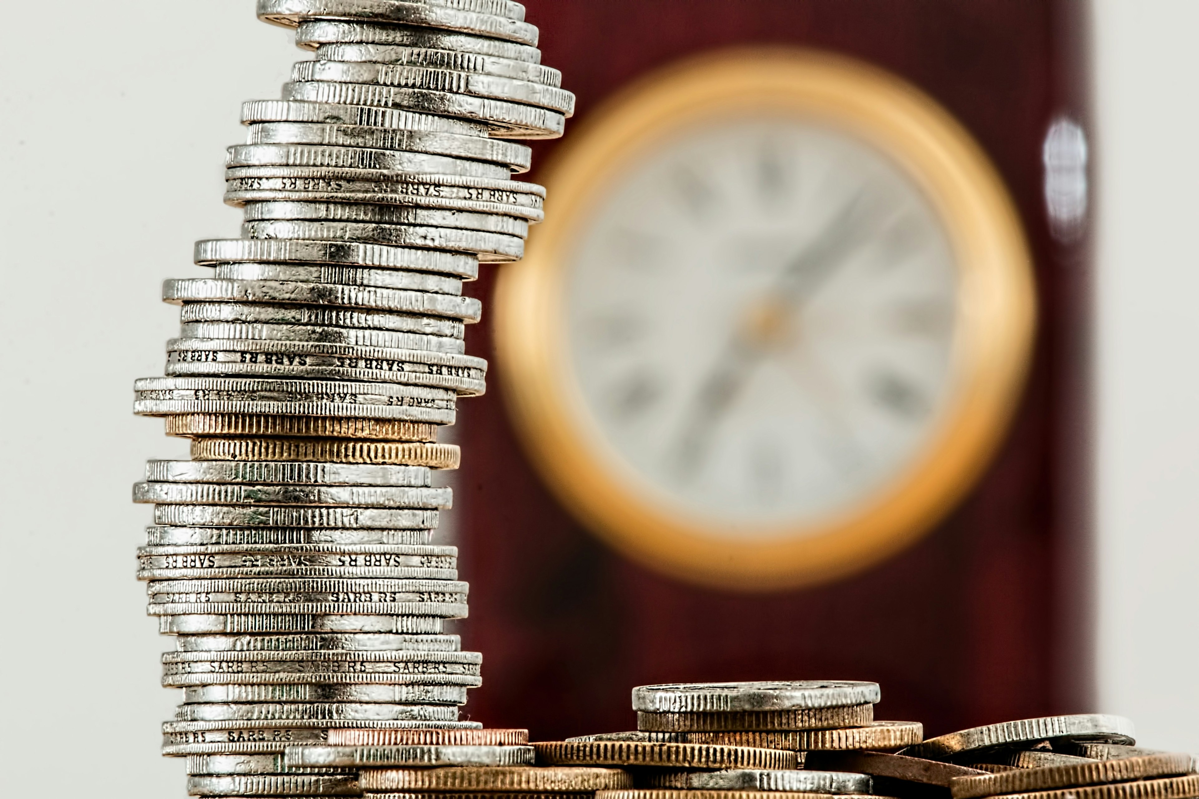 A stack of coins in the foreground with an out-of-focus clock in the background representing "time is money"
