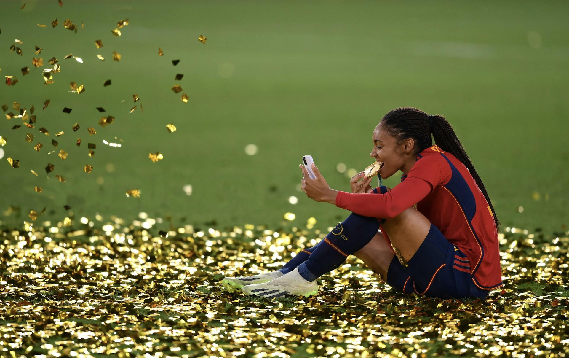 A woman in a pile of confetti, bitting a medal and taking a selfie. 