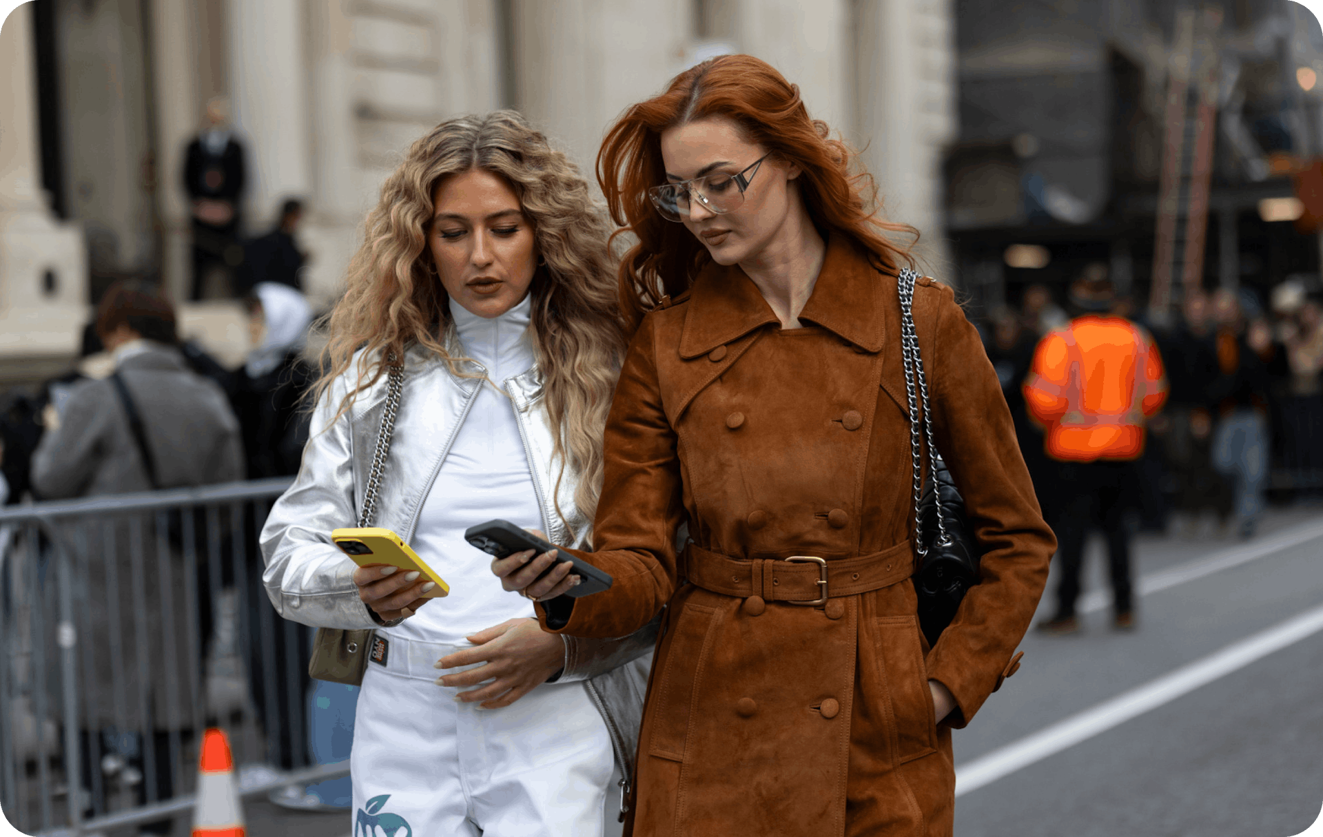 Street Style photograph in which two girls with long hair and winter clothes walk around smiling and looking at their mobile phone screens.