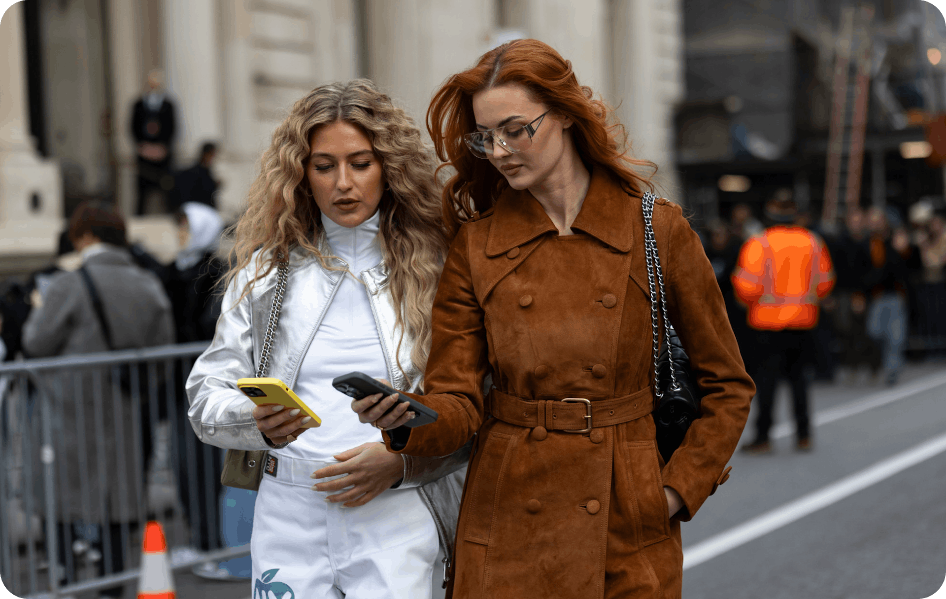 Street Style photograph in which two girls with long hair and winter clothes walk around smiling and looking at their mobile phone screens. 