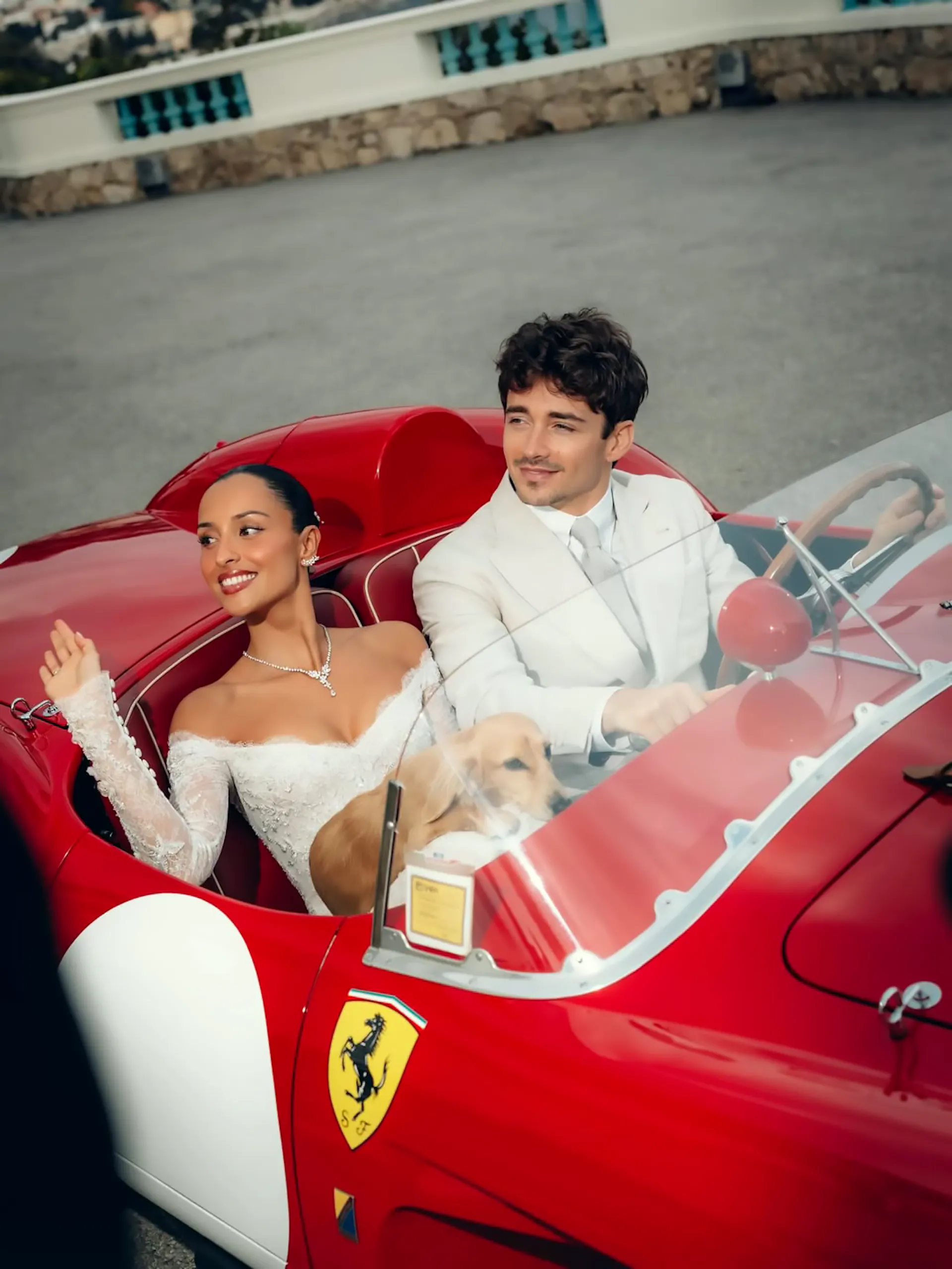 A high-angle photograph of Formula 1 driver Charles Leclerc in a white suit and his wife Alexandra Saint Mleux in a white lace wedding gown, driving a rare vintage Ferrari 250 Testa Rossa. Their miniature dachshund puppy, Leo, sits between them in the cockpit as Alexandra smiles and waves.