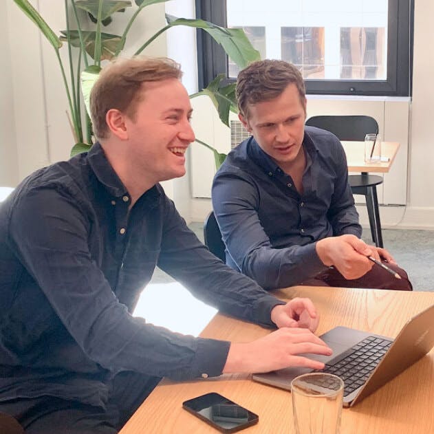 Two people sat working at a laptop, one is laughing and the other is pointing at the screen