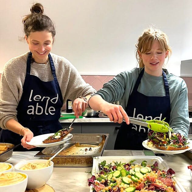 Two people wearing Lendable branded aprons dishing food on to plates from a large bowl of salad