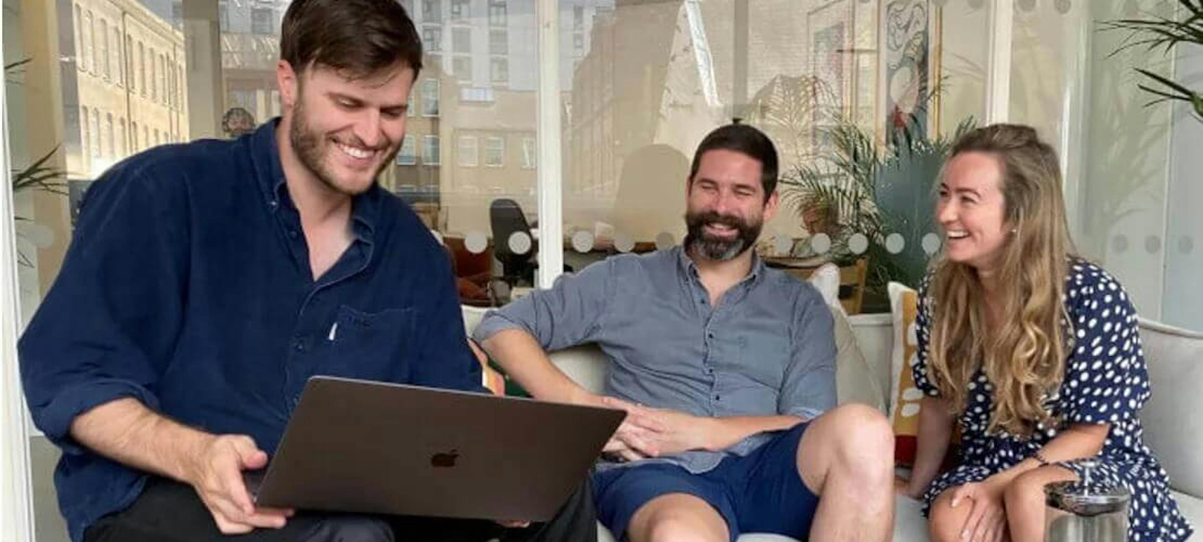 A team of three people sat on a sofa smiling whilst looking at a laptop