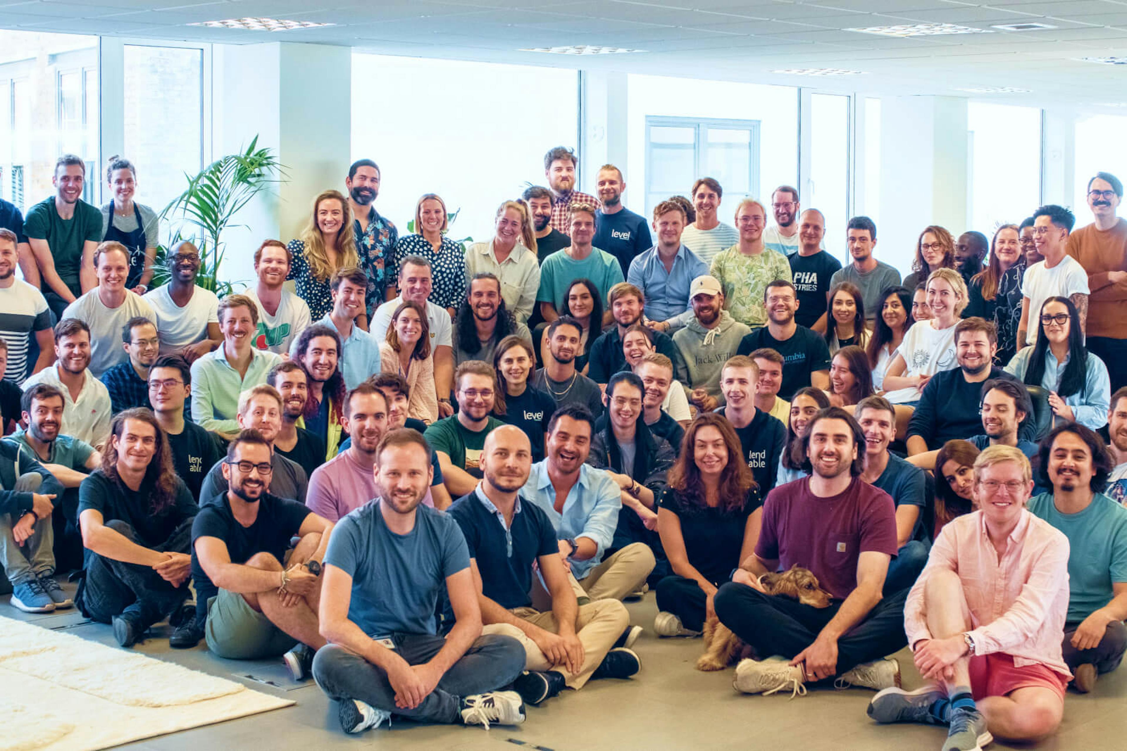 A large group of around 50 people, and one dog, in an office facing the camera and smiling