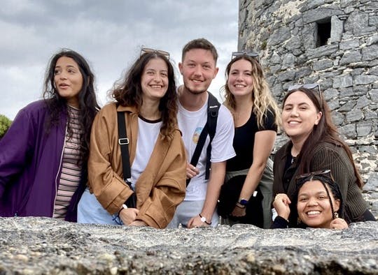 A group of six smiling young adults standing behind a stone wall with a tall, historic stone castle tower in the background under a cloudy sky.