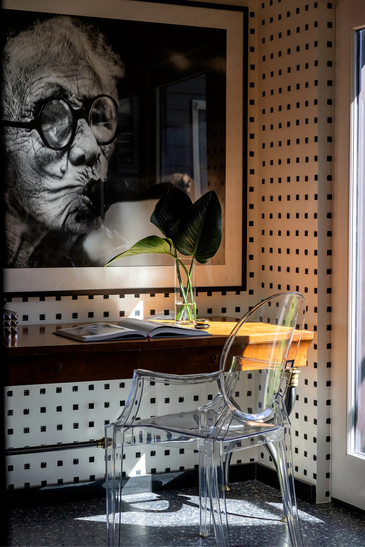 Interior design workspace with patterned walls, framed portrait artwork, wood desk, and transparent chair in natural light