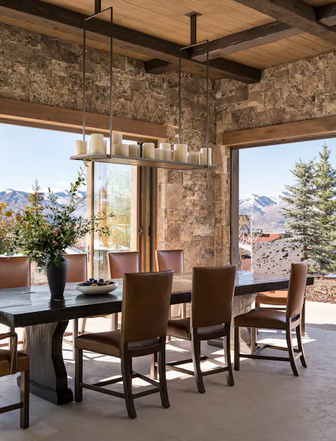 Rustic luxury dining room with wood table, leather chairs, stone walls, and mountain views through large windows