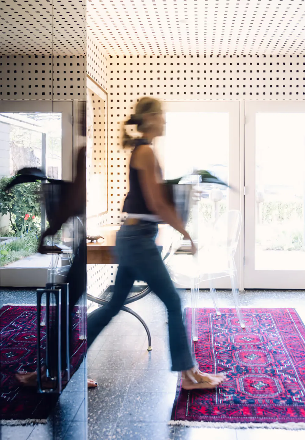 Leslie Schofield walking through patterned interior carrying plant in modern space