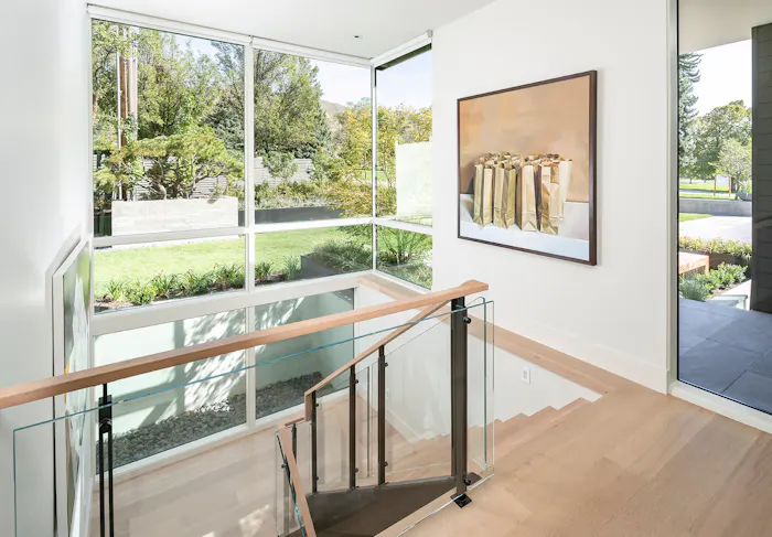 Modern staircase landing with floor-to-ceiling corner windows, glass railing, and contemporary artwork by Leslie Schofield Studio