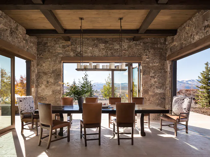 Luxury mountain dining room with stone walls, exposed wood beam ceiling, and panoramic landscape views by Leslie Schofield Studio