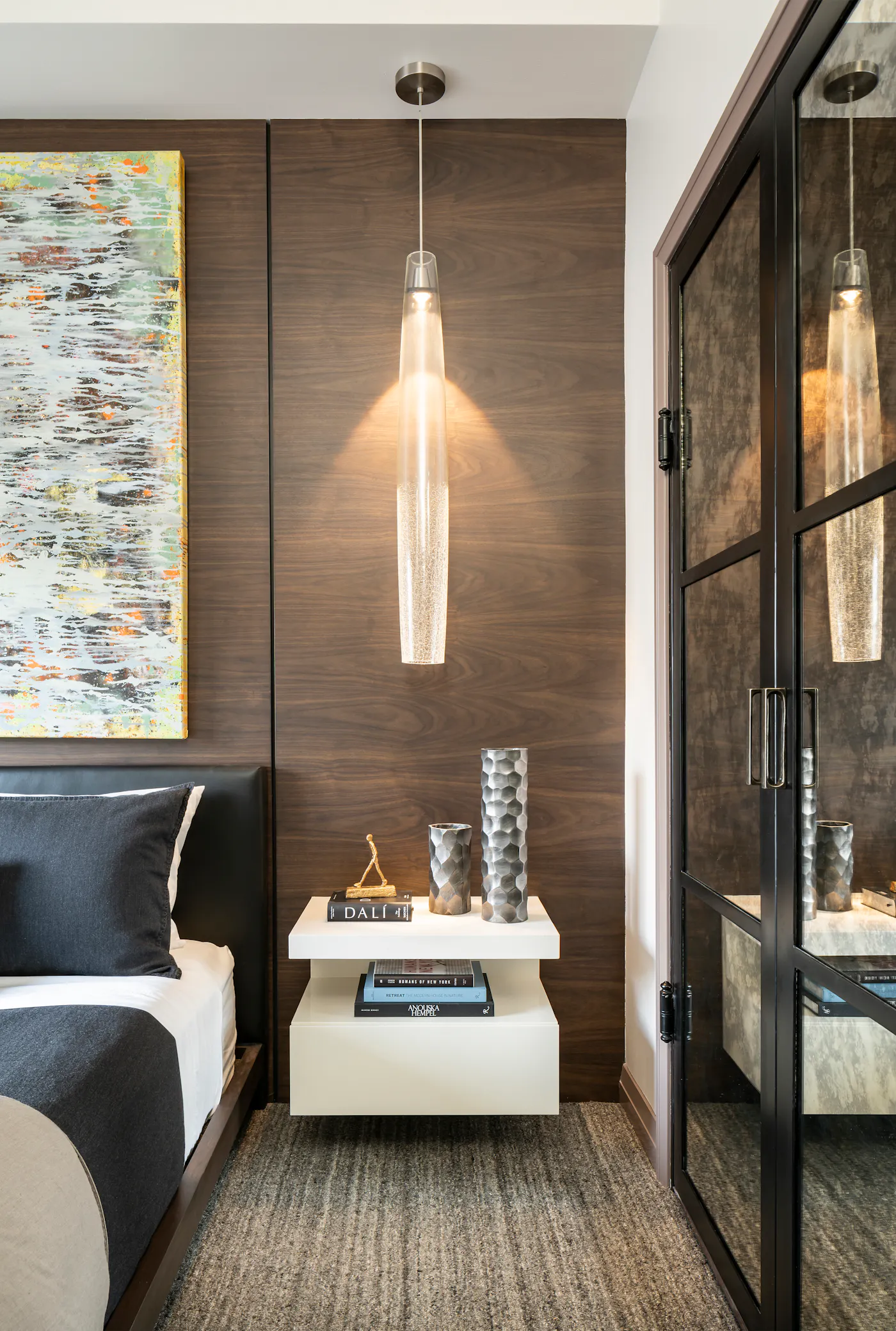 Modern luxury bedroom vignette featuring a floating nightstand, walnut wall paneling, sculptural glass pendant light, custom steel-framed glass doors, and contemporary designer décor.