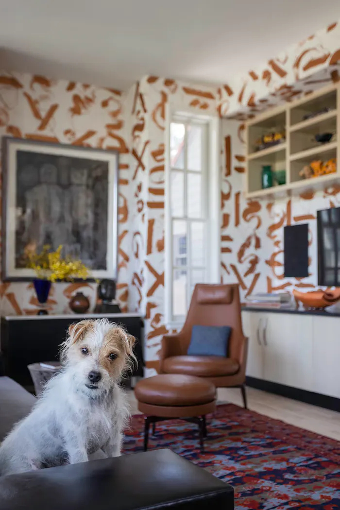 Modern residential office with artistic wallpaper, layered decor accents, warm wood tones, and a small terrier sitting in the foreground