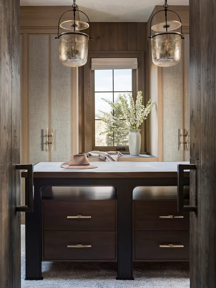 Luxury custom walk-in closet featuring dark wood cabinetry, linen cabinet panels, oversized pendant lighting, and a central storage island