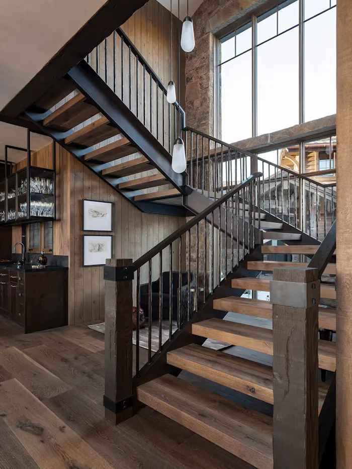 Modern rustic staircase design with exposed wood textures, custom metal railings, and dramatic natural light in a luxury mountain residence