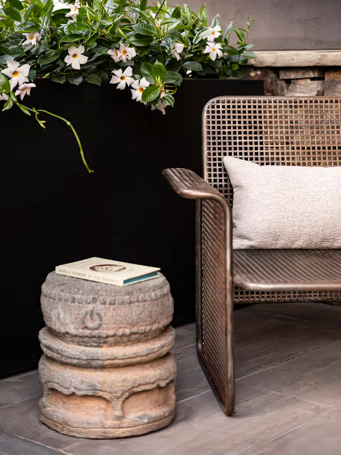 Stylish exterior patio corner with a textured metal lounge chair, sculptural stone side table, and lush greenery in a modern residential landscape