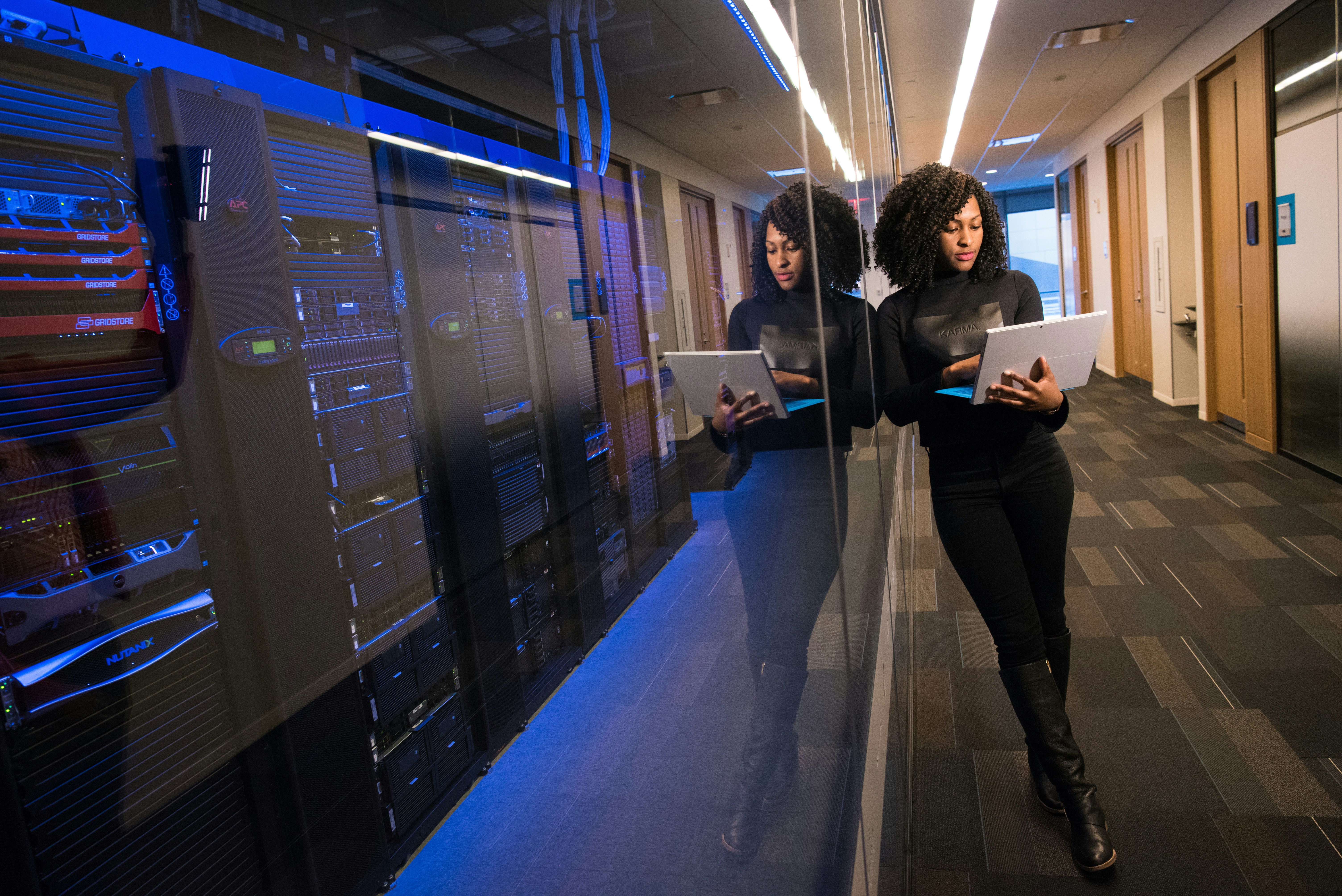 A woman using her laptop leaning on a window to a dark server room