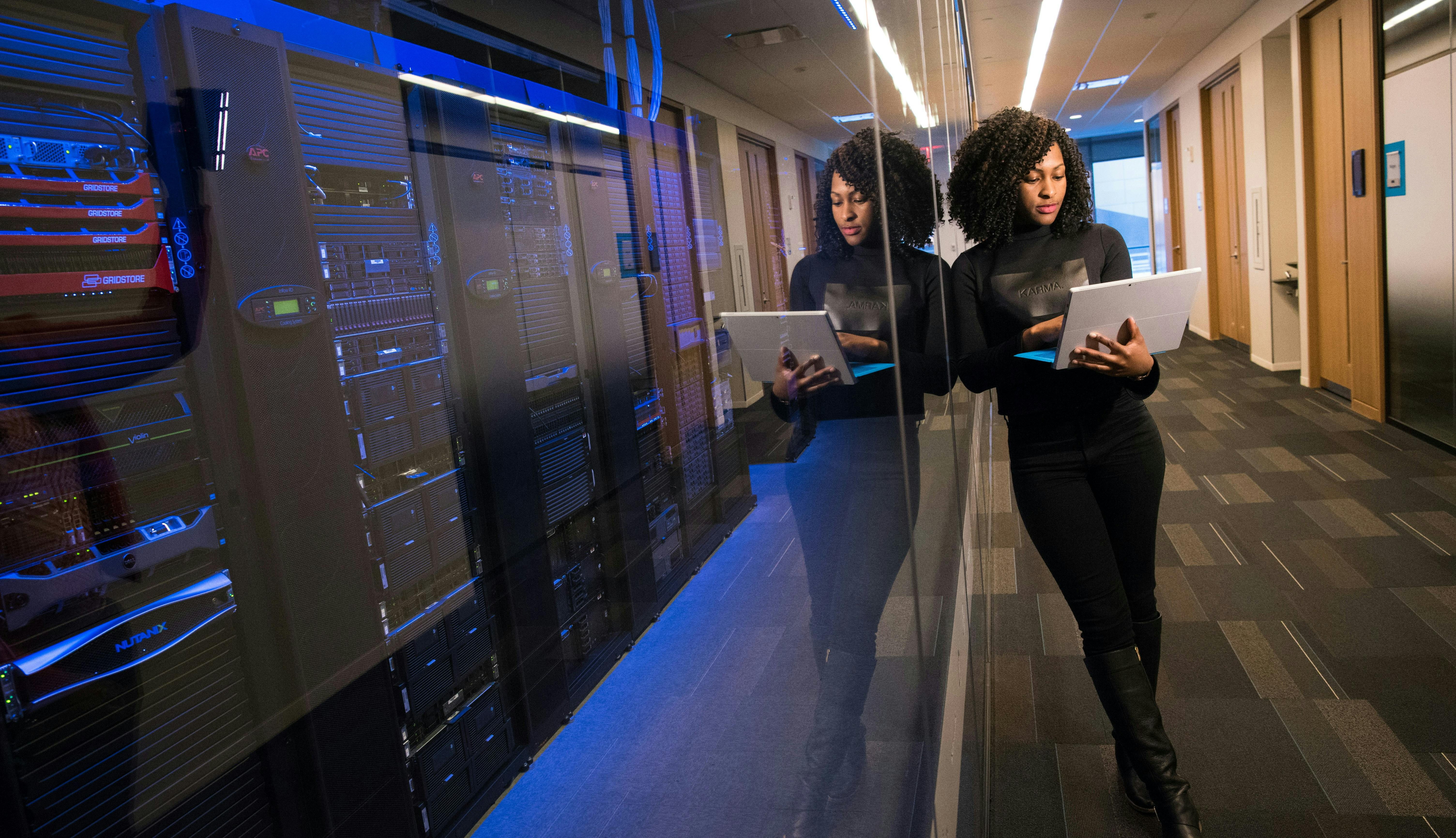 A woman using her laptop leaning on a window to a dark server room