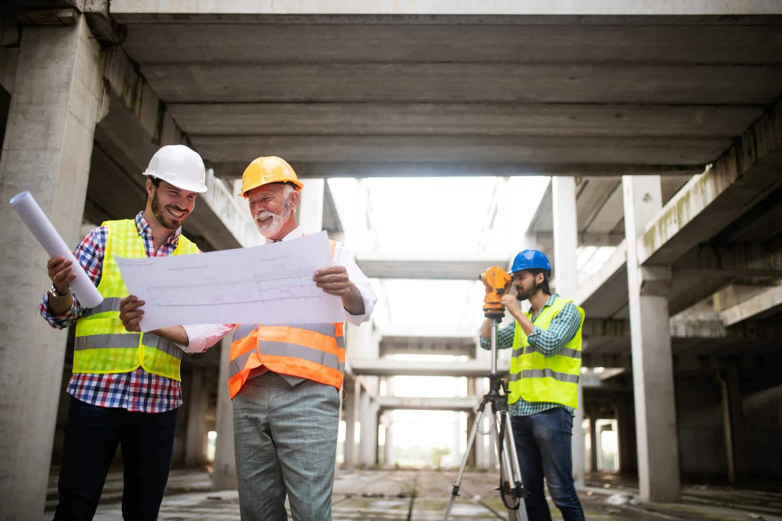 2 construction engineers at a construction site looking at a schematic and smiling while a surveyor works in the background.