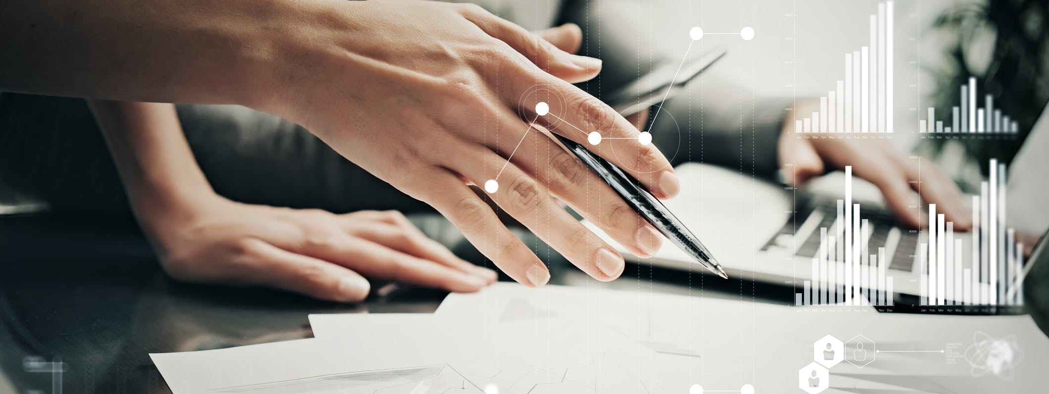 A hand holding a pen above sheets of paper. Stats and growth icons on screen.