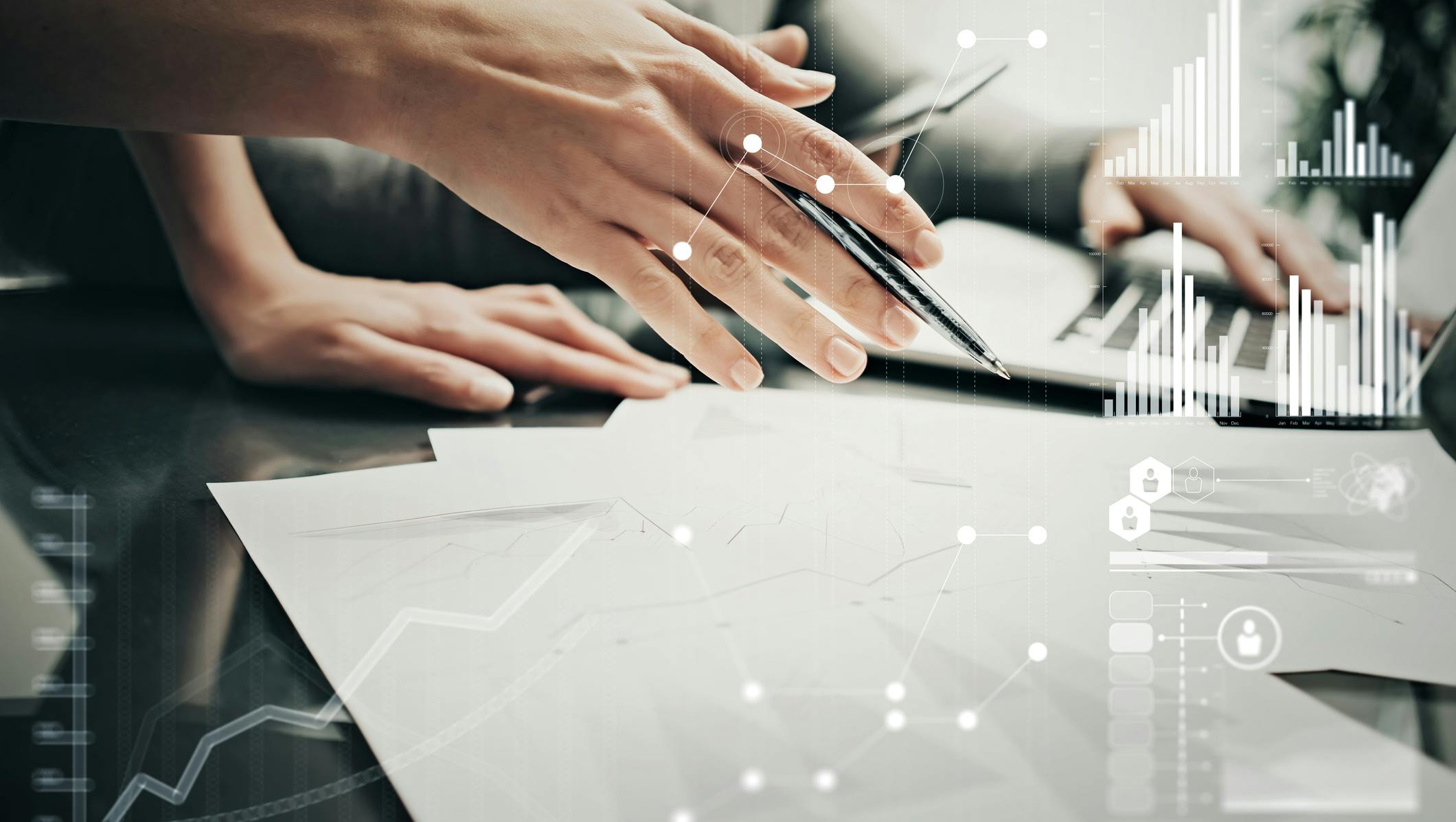 A hand holding a pen over paper. Stats and growth icons on screen.