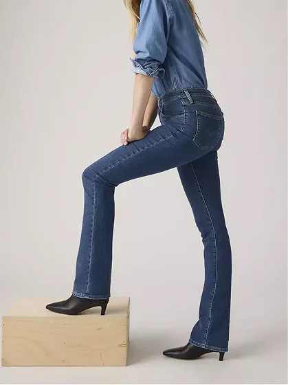 Woman in medium-wash bootcut jeans and denim shirt standing on a wooden block.