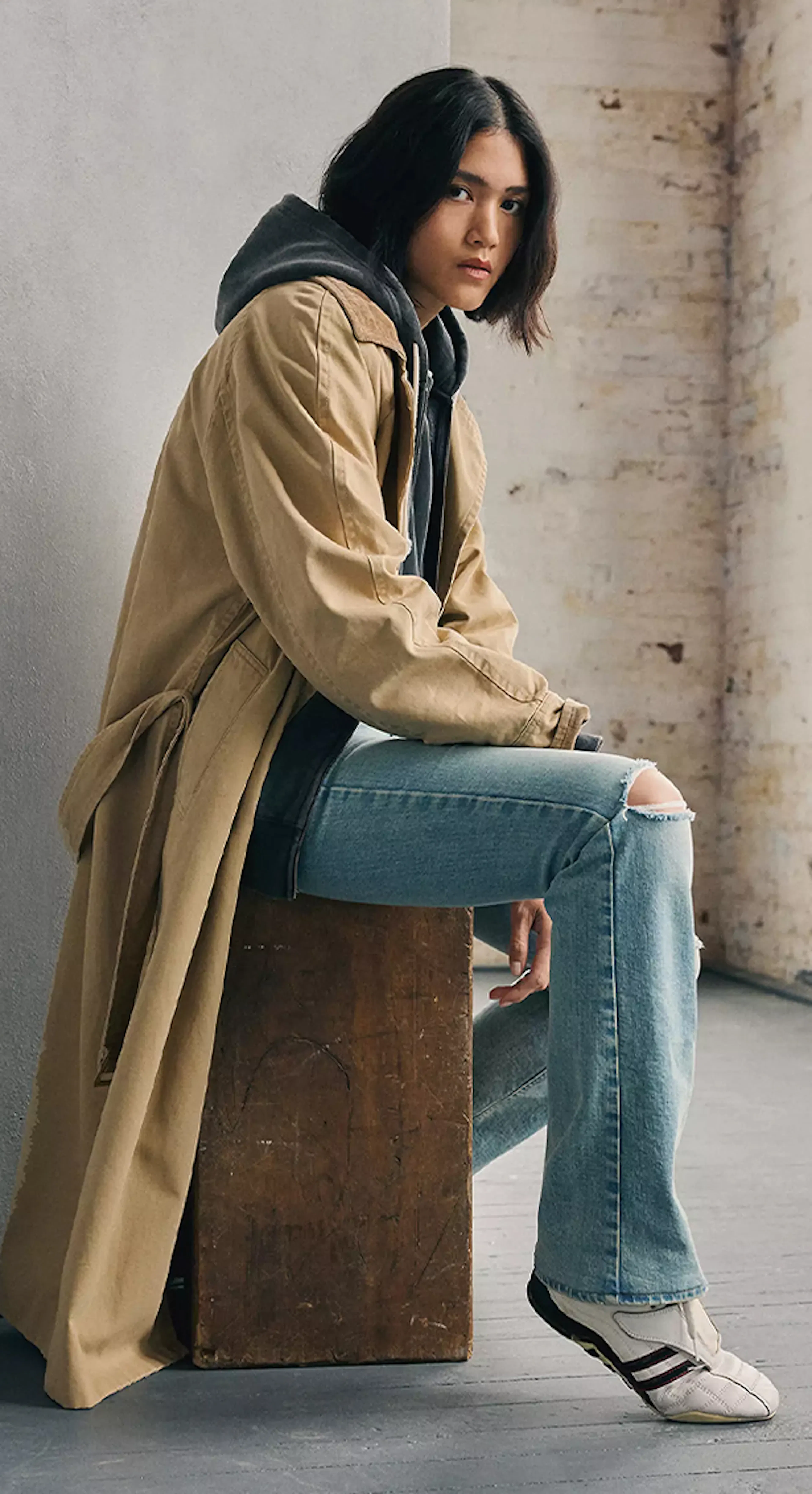 Woman wearing light-wash jeans, white sneakers, and a tan trench coat layered over a hoodie, sitting casually on a wooden block.