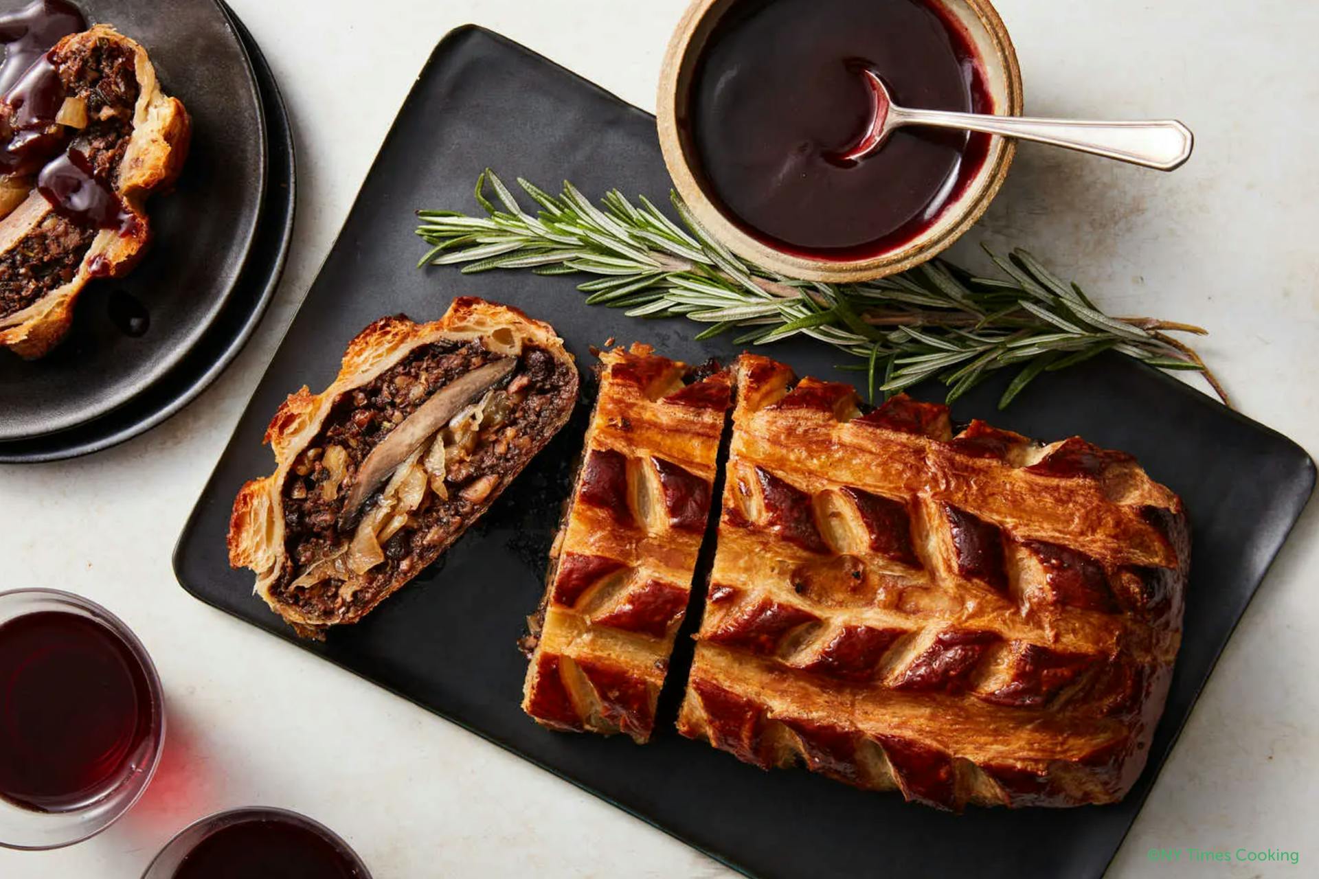 Een gedekte tafel met zwart bord en daarop een bladerdeeggerecht: Vegetarische champignon Wellington en een takje Rozemarijn en een potjes saus.