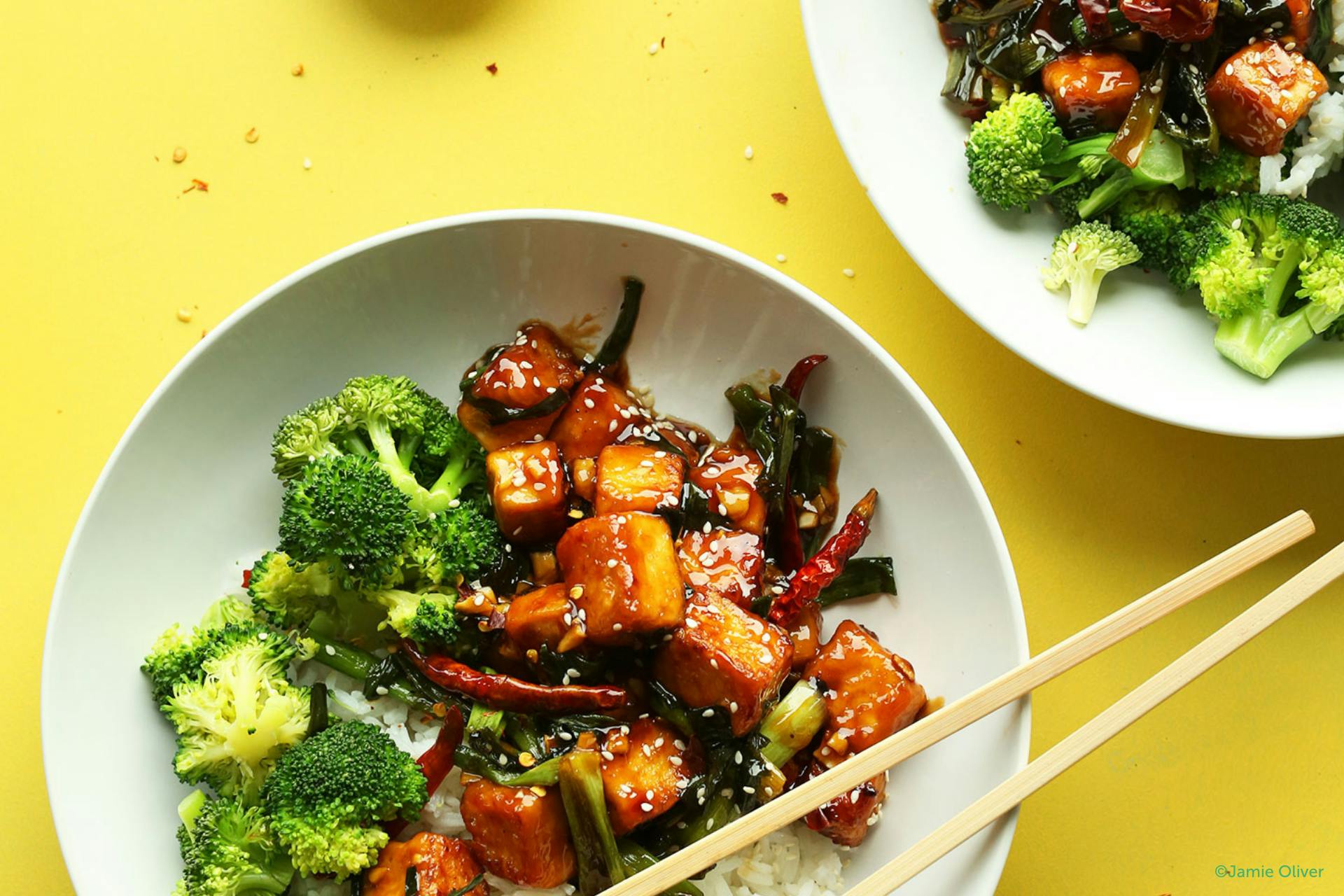 Foto met 2 kommen gevuld met General Tso's tofu, broccoli en chopsticks op een gele achtergrond