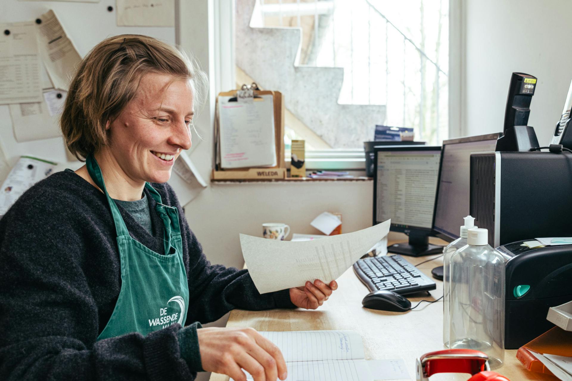 Hilde met haar winkelschort aan, aan een bureau omringd door papier en schermen