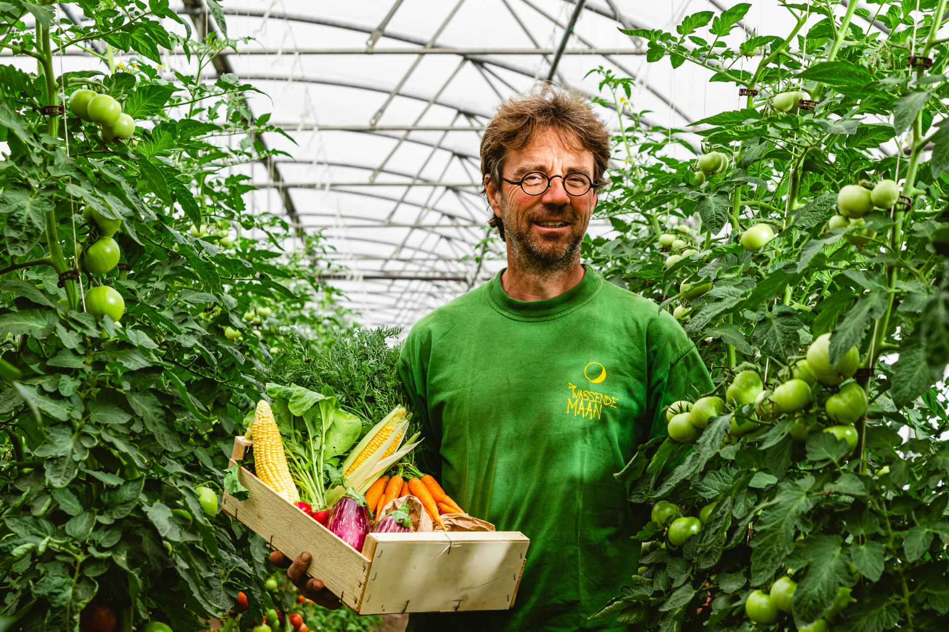 Stefan in de serres tussen de tomatenplanten die een houten kistje met vers geoogste groenten vasthoudt