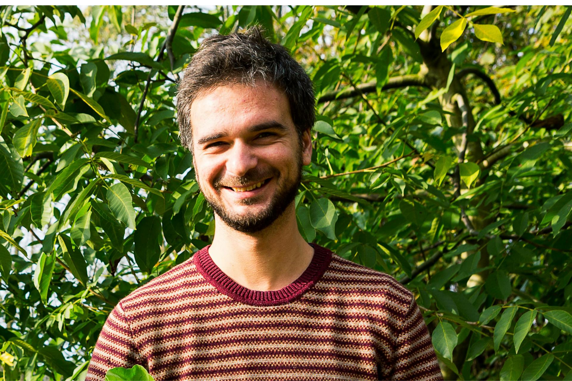 Headshot van een lachende Niels buiten, met zijn hoofd tussen de blaadjes van de bomen