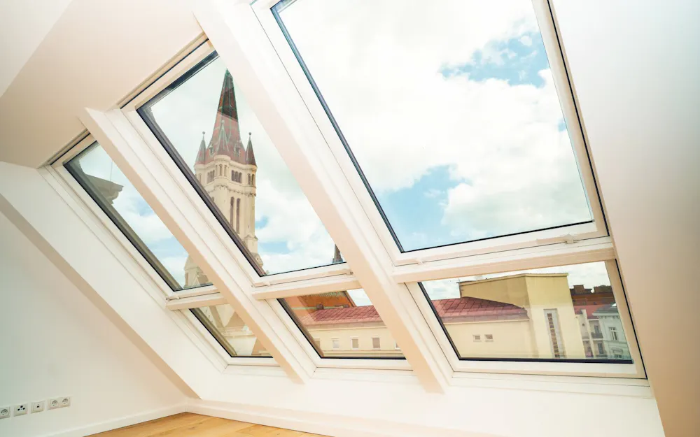 Zwei geöffnete Fakro Schwingfenster in einem hellen Dachgeschoss mit Blick auf Kirchturm und Dächer Zwei geöffnete Fakro Schwingfenster in einem hellen Dachgeschoss mit Blick auf Kirchturm und Dächer
