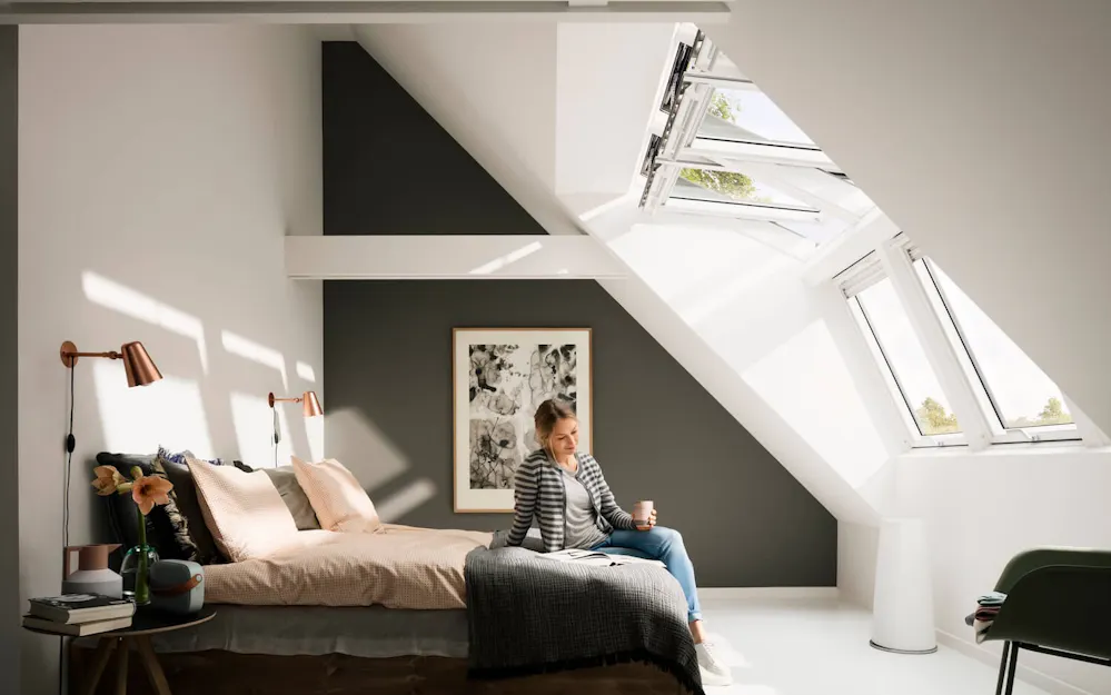Frau sitzt auf dem Bett in einem hellen Schlafzimmer mit Velux Dachfenstern Frau sitzt auf dem Bett in einem hellen Schlafzimmer mit Velux Dachfenstern