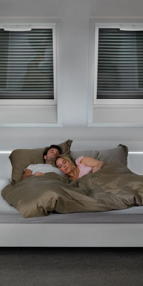 Paar schläft in einem Bett unter zwei Roto Dachfenstern mit Sonnenschutzrollo Paar schläft in einem Bett unter zwei Roto Dachfenstern mit Sonnenschutzrollo
