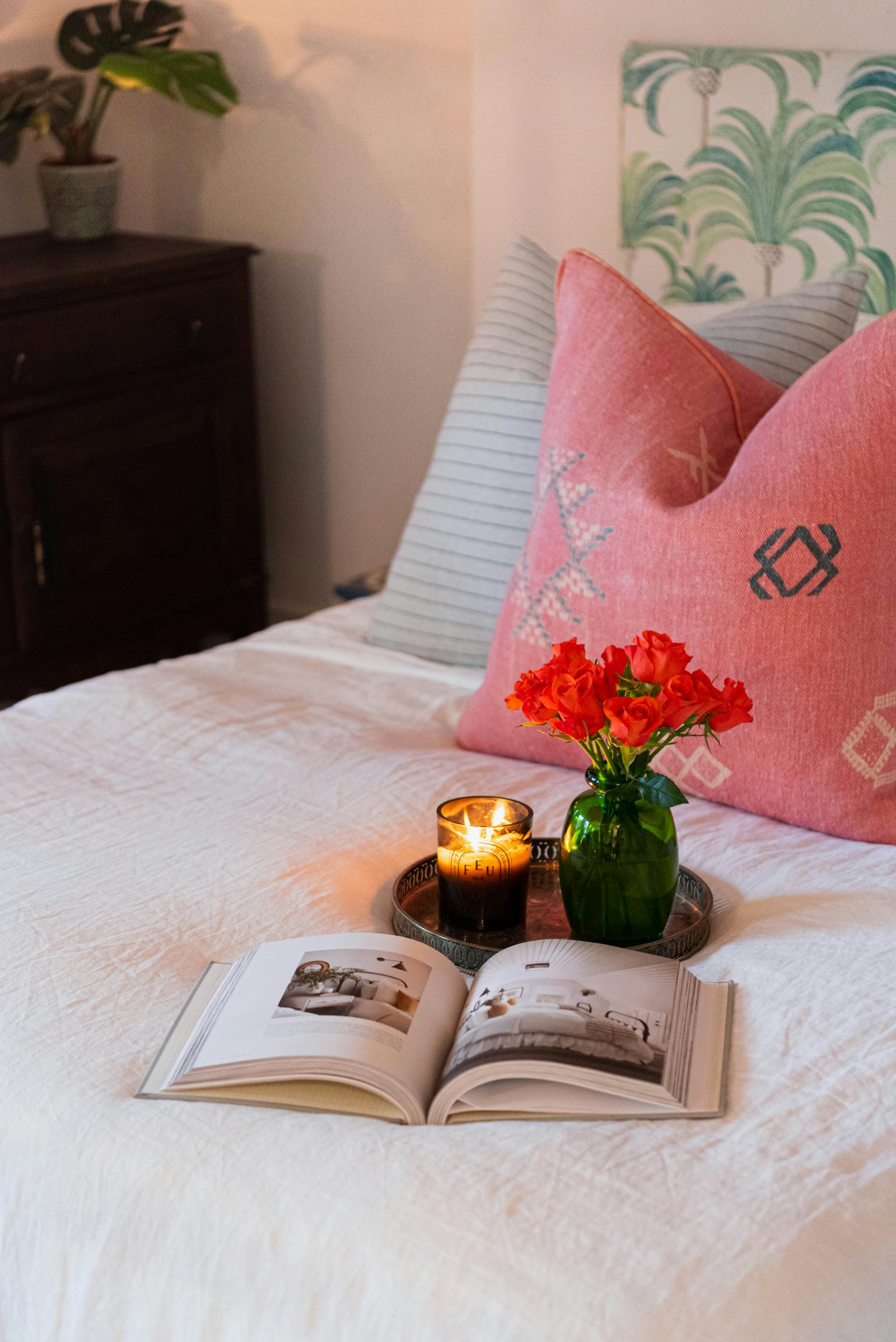 Close up image of flowers and a book on a bed that has pink and blue cushions and a palm tree pattered headboard