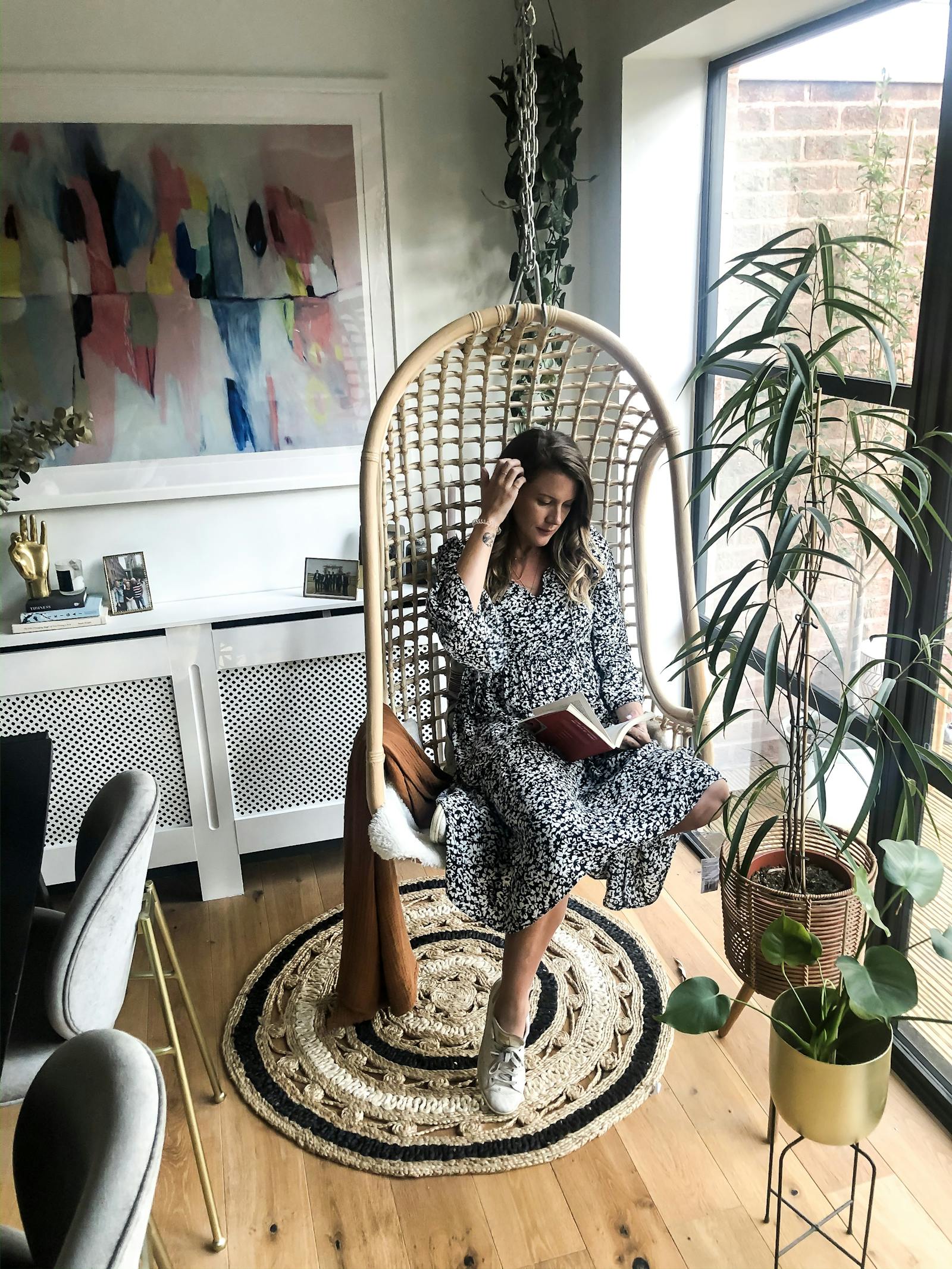 A woman sitting in a rattan chair swing and reading a book, with an abstract colourful painting behind her