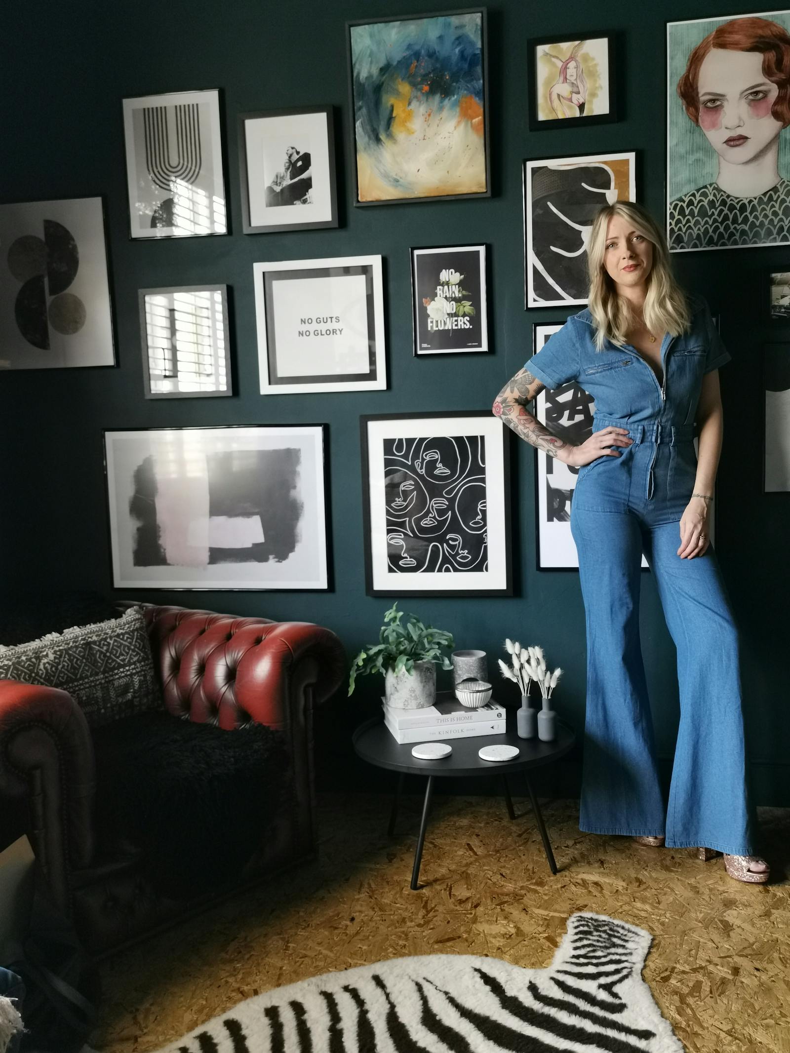 Blonde woman in a denim jumpsuit posing in front of a gallery wall painted in dark teal