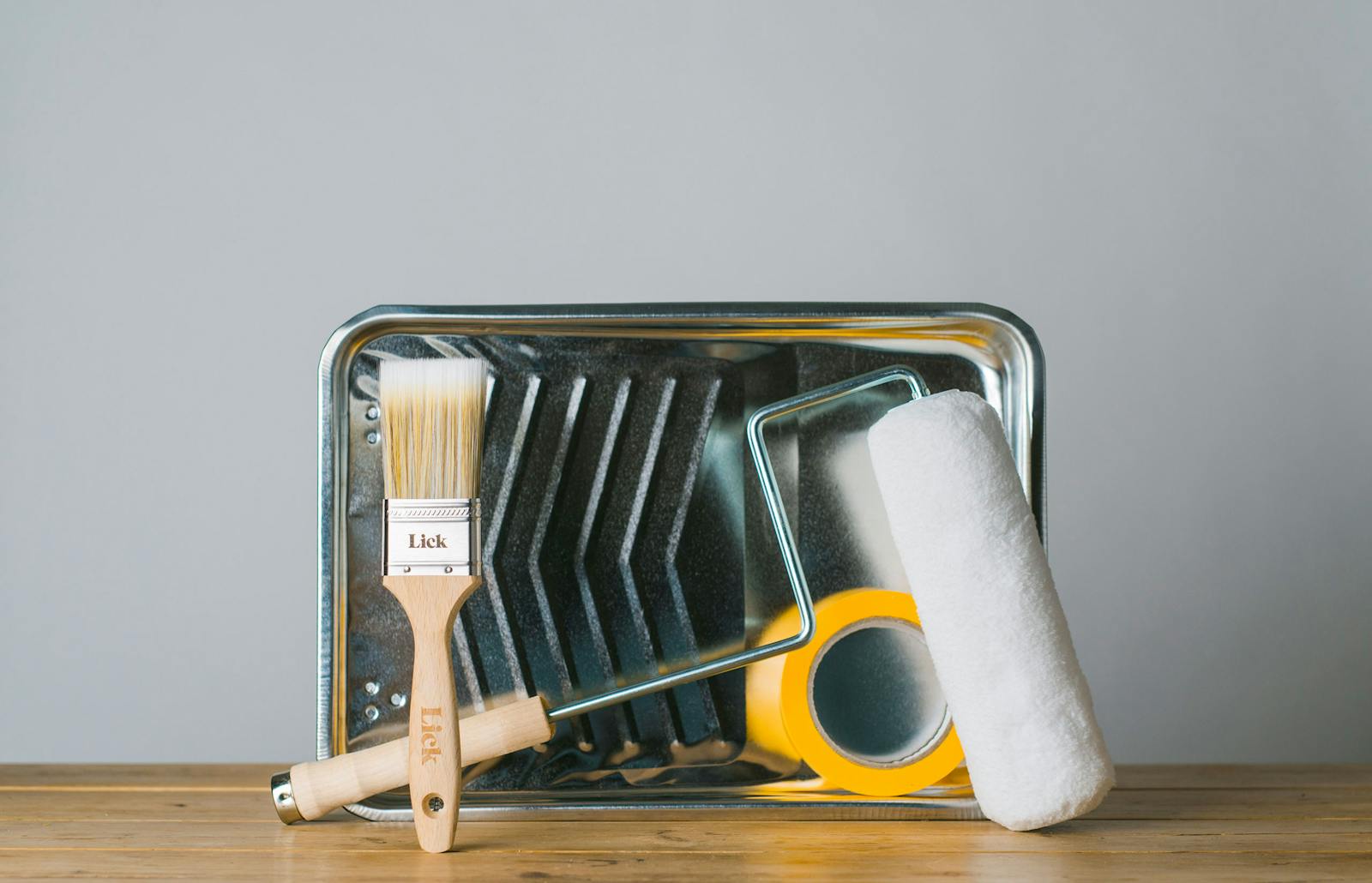 An essential 5 peice collection of Lick painting and decorating tools including yellow decorating tape, a 2inch wooden handle paintbrush with natural hairs, a 9inch wooden handle high-quality roller with fluffy white roller head and a metal paint tray.