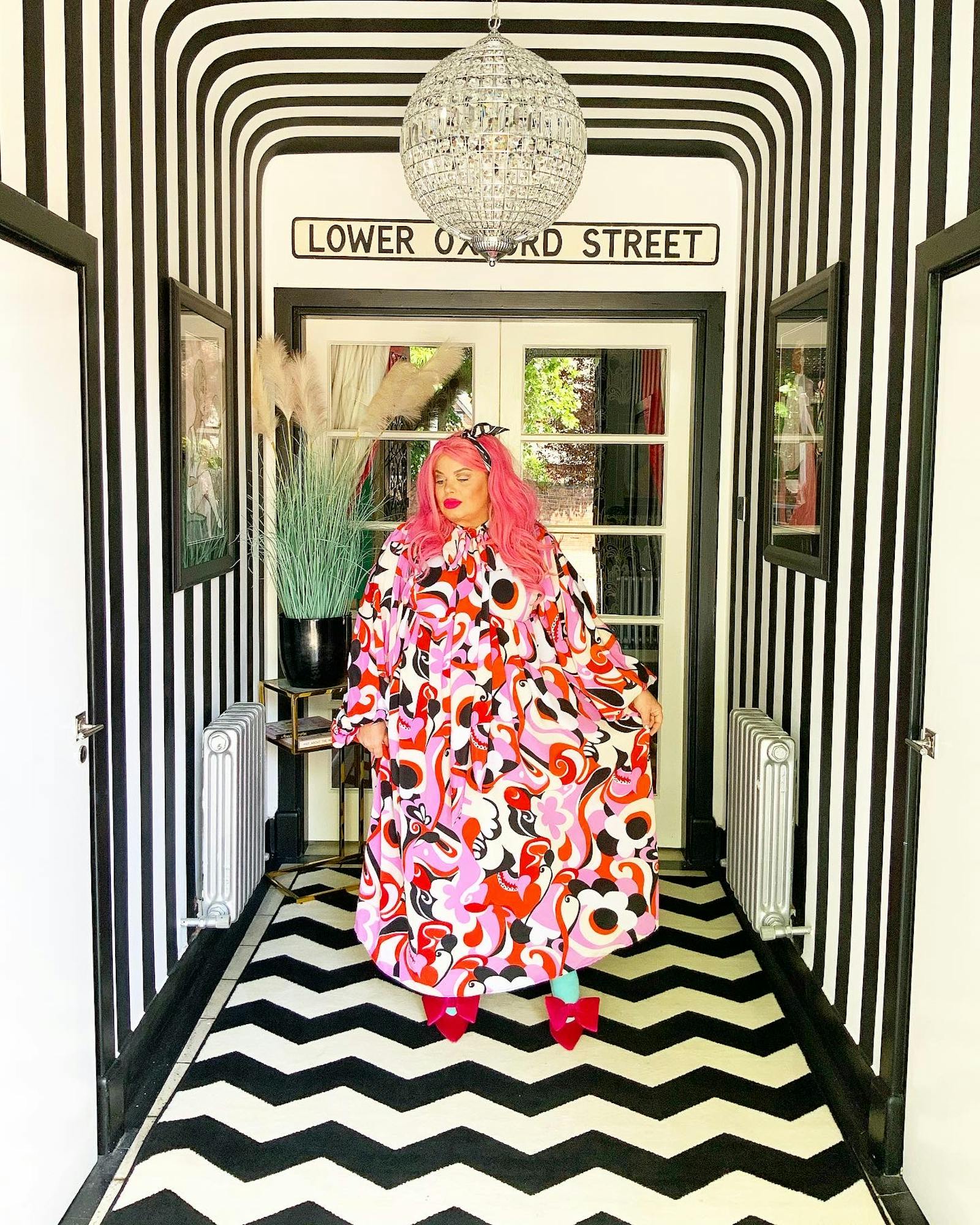 woman with pink hair and patterned dress posing in the middle of a hallway covered in stripes