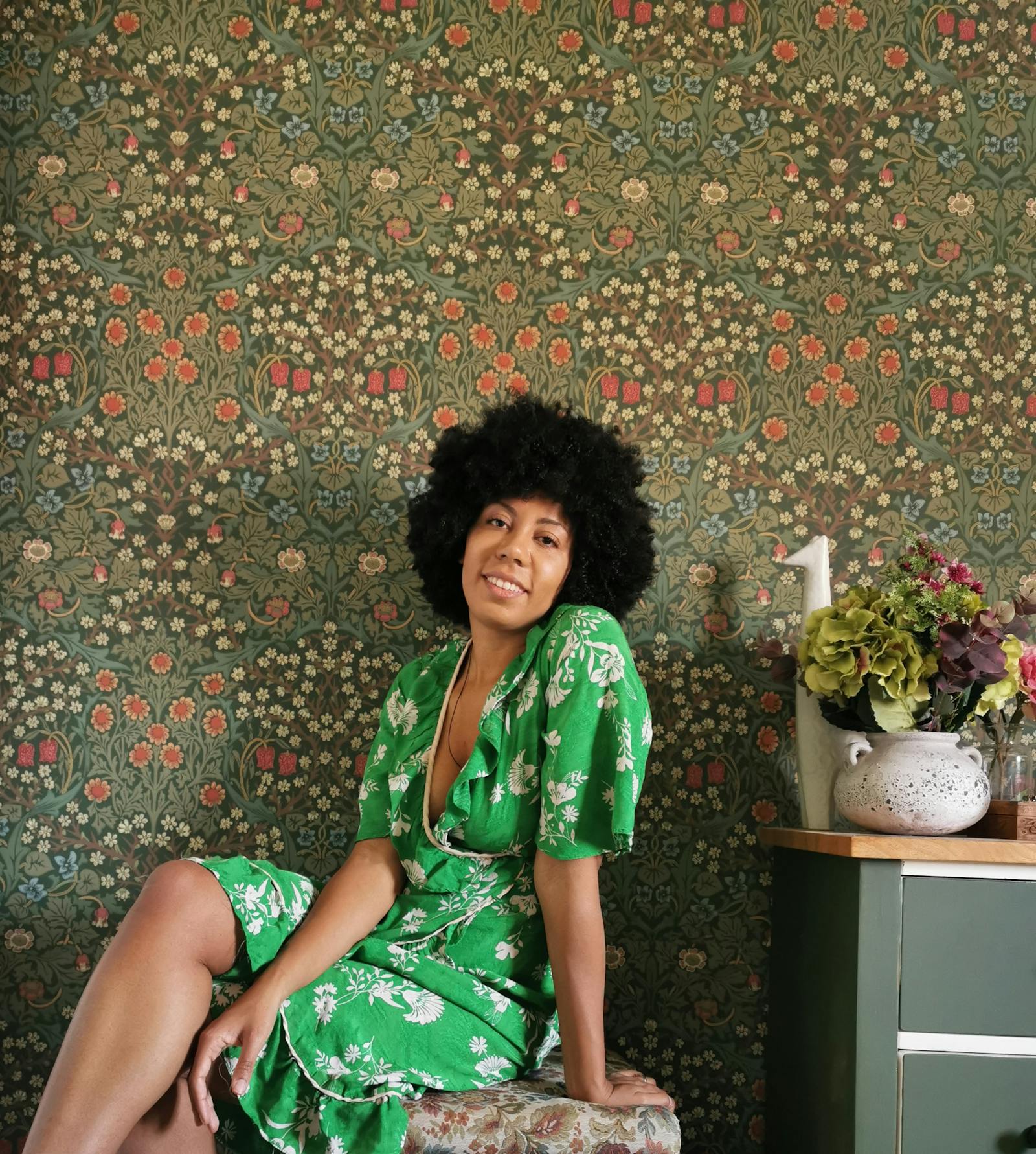 Woman in a floral green dress sitting in front of a grass green floral patterned wallpaper