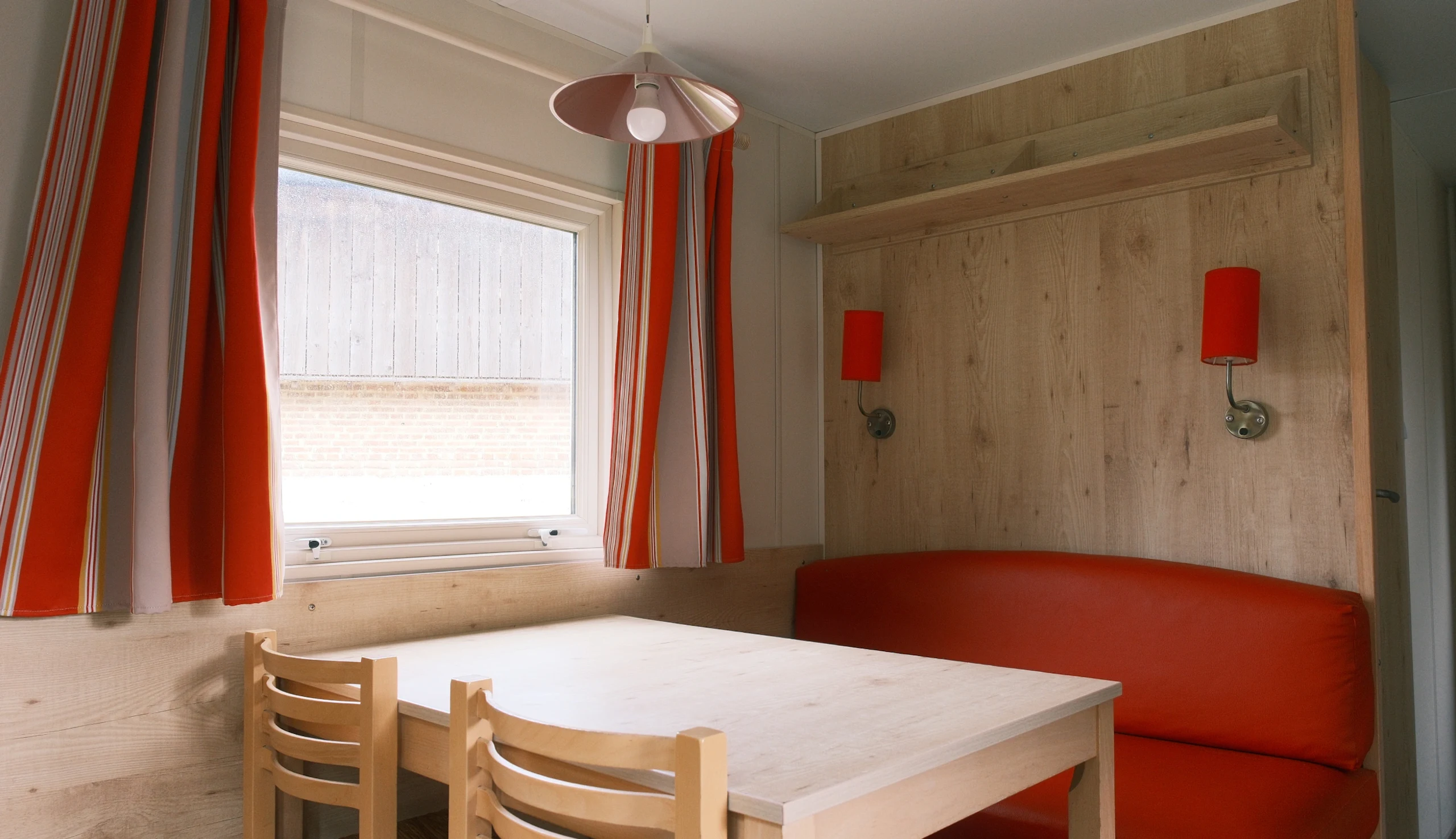 Table et chaises en bois, banquette rouge, appliques au mur et rideaux rouges et beiges