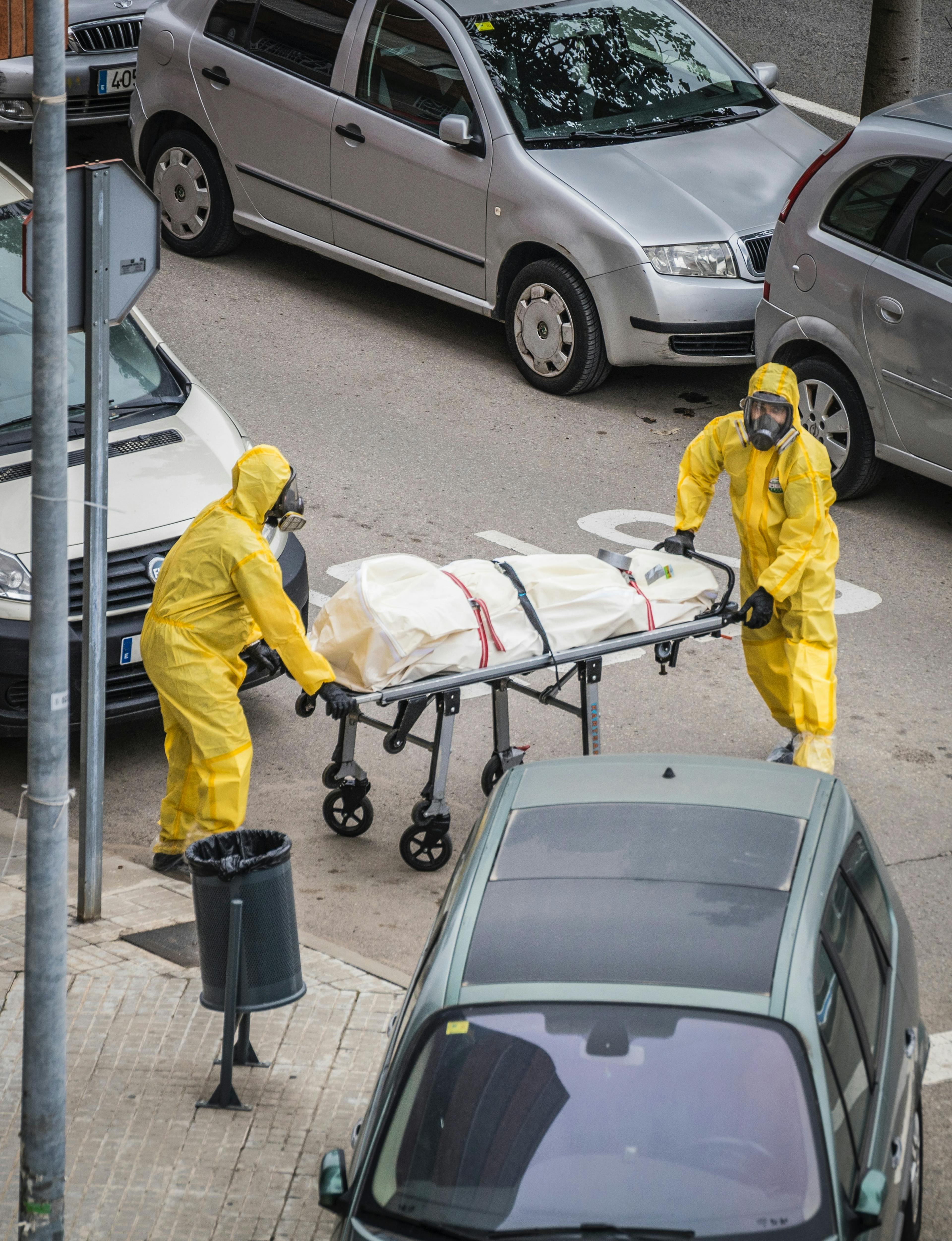 Two people in yellow PPE wheeling wrapped body through street